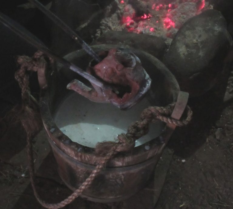 A hot clay teapot being dipped into a bucket of milk beside glowing embers.