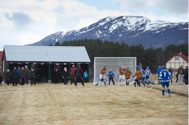 A shinty match taking place on a field beside the Boleskine pavilion, with snow-covered hills behind.