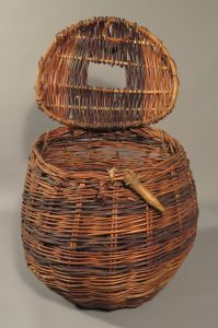 A large round wicker basket with a hinged lid that is open, showing a square opening in the lid.