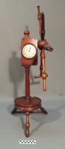 A tall wooden yarn winder with a round base, upright post and attached clock‑like gauge.