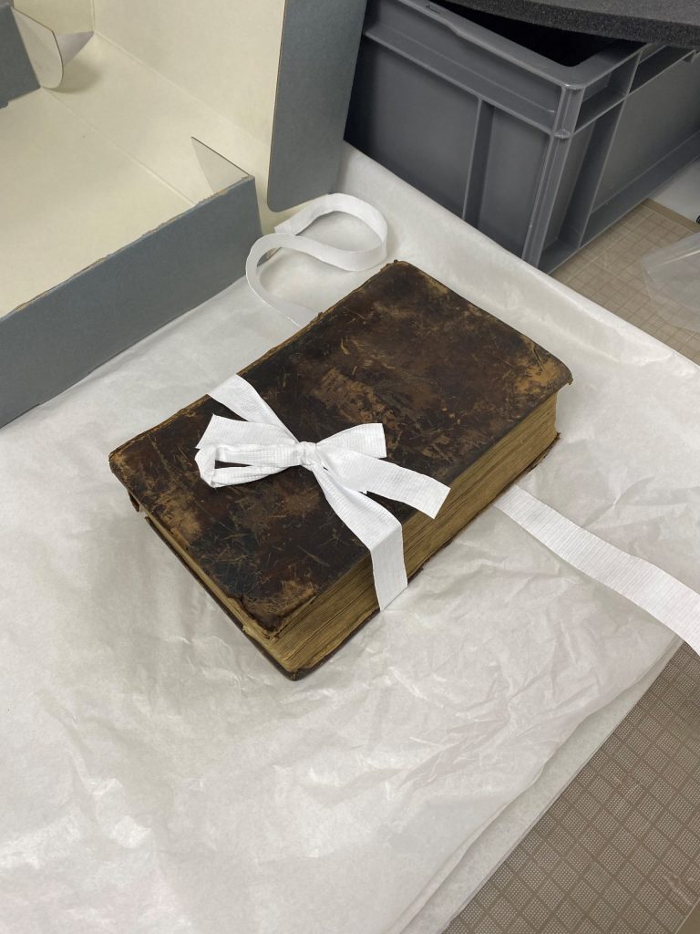 An old, worn book tied with a white fabric ribbon, resting on white tissue paper for support.