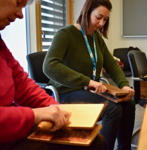 Two people sitting and carding wool using wooden carders.