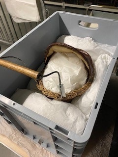 A wicker-handled wooden pram packed inside a plastic crate, cushioned with white tissue paper.