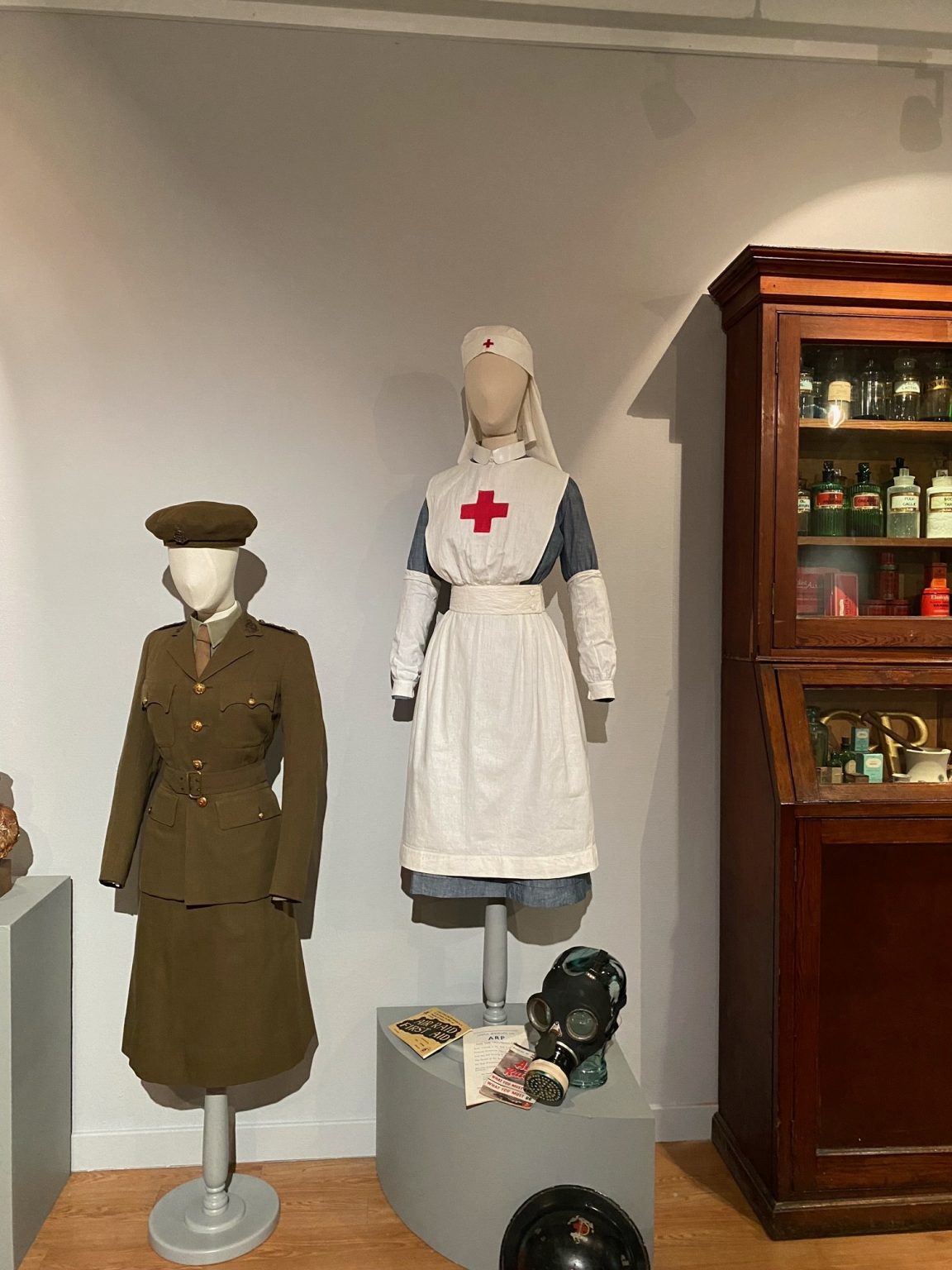 Two mannequins dressed in a military uniform and a nurse’s uniform, displayed beside a wooden cabinet of medical artefacts.