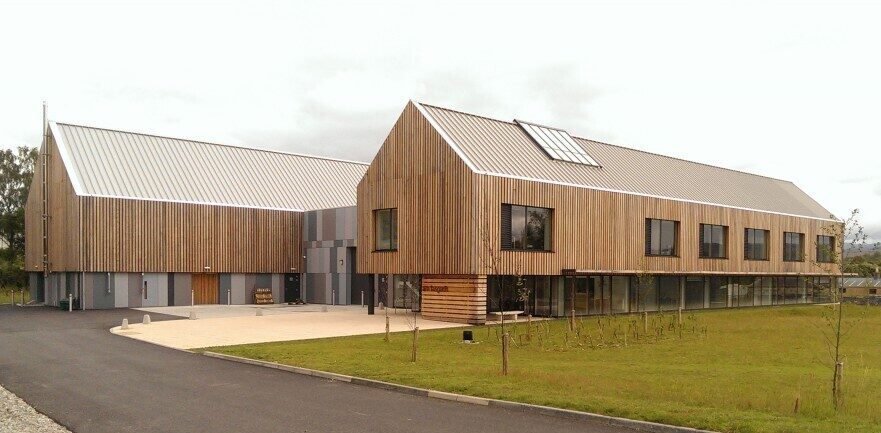 Modern wooden-clad building with sloped metal roof, large windows, and paved driveway on grassy grounds.
