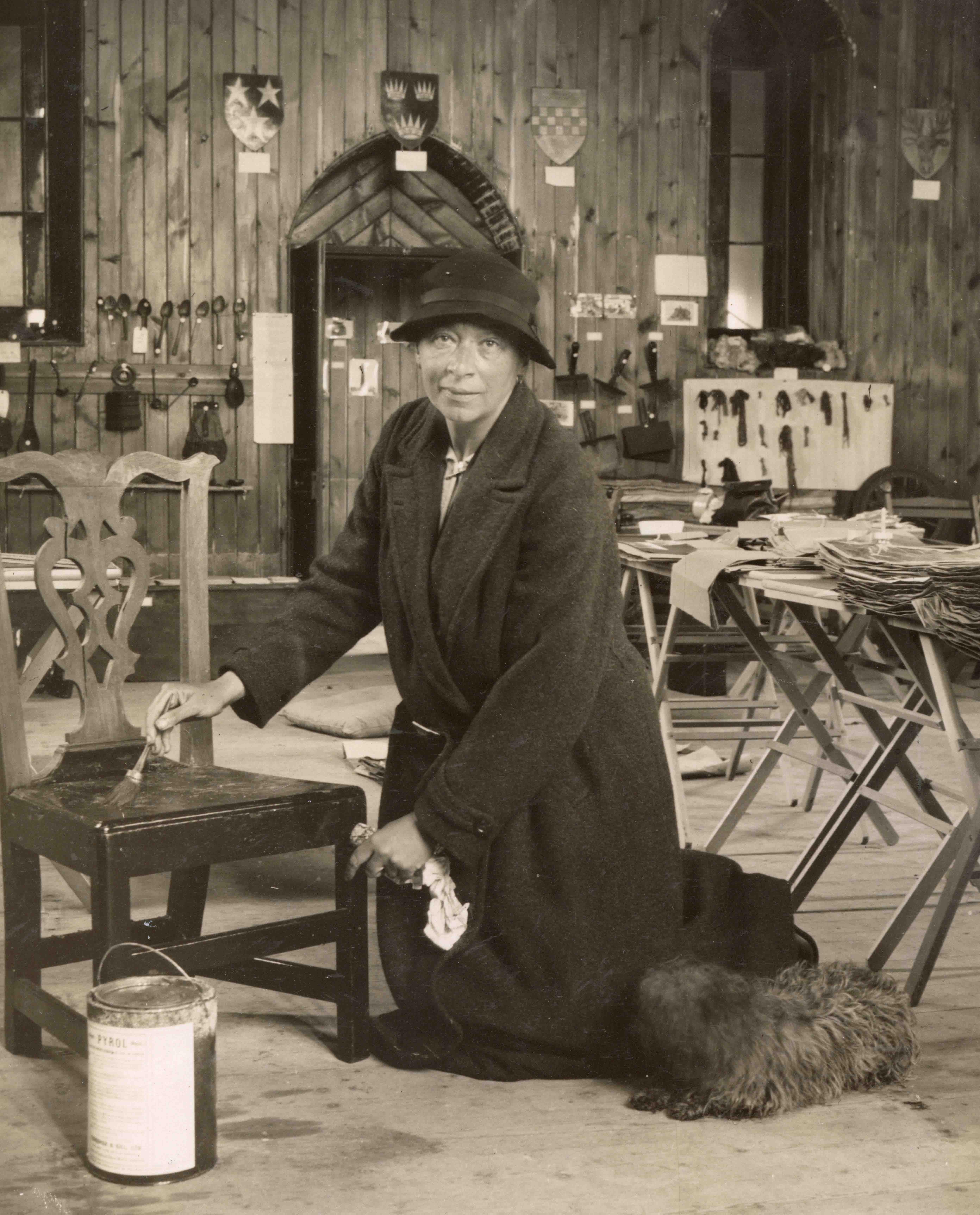 Black and white image of woman kneeling on floor in a wood panelled room, painting a wooden chair with woodworm treatment. She is looking directly at the camera, and wearing a hat and long coat. There is a small dog sitting next to her. In the background there are many artefacts and museum exhibits on display.