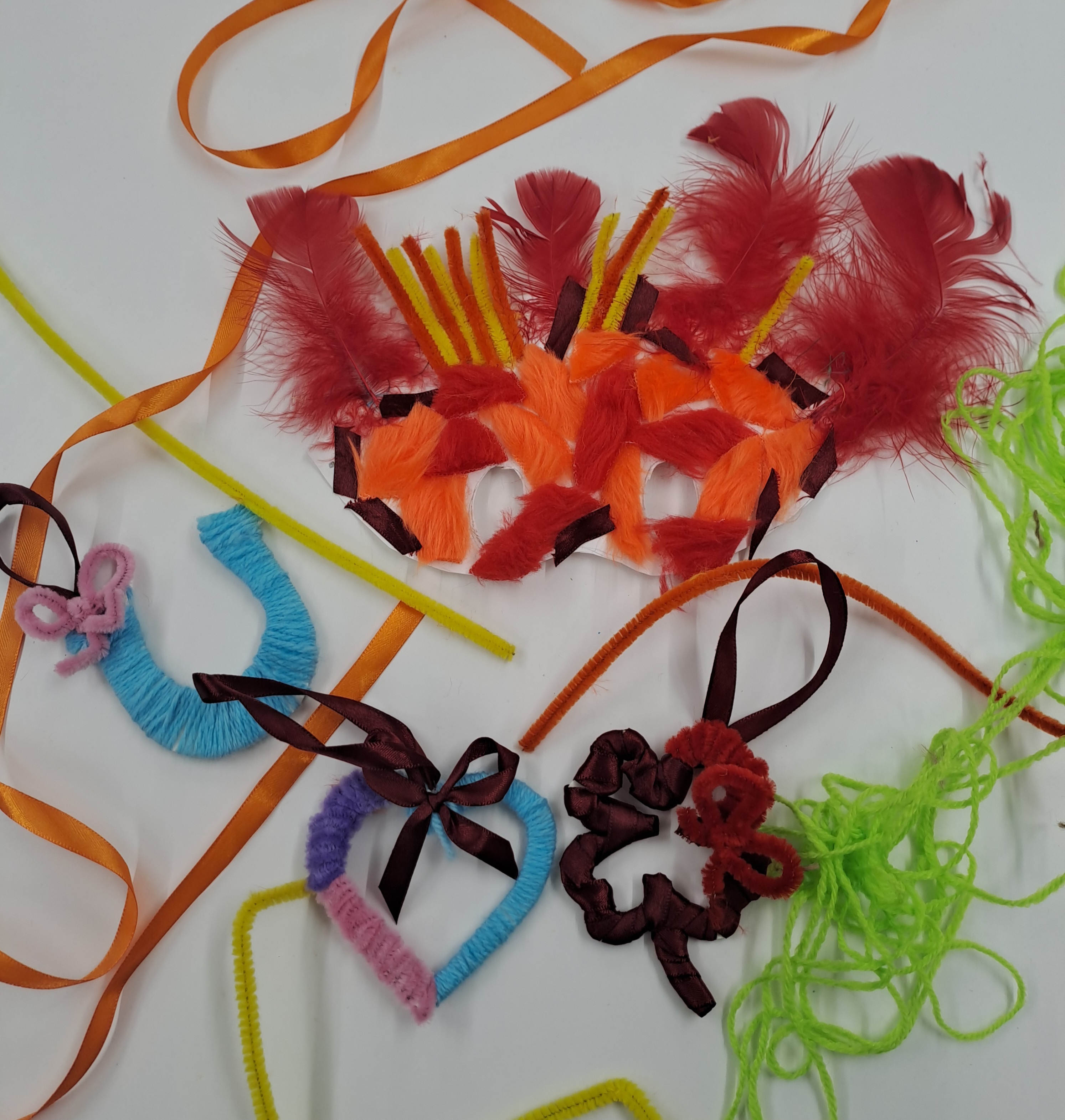 Colourful craft shapes made from feathers, pipe cleaners, ribbon, and yarn arranged on a white background.