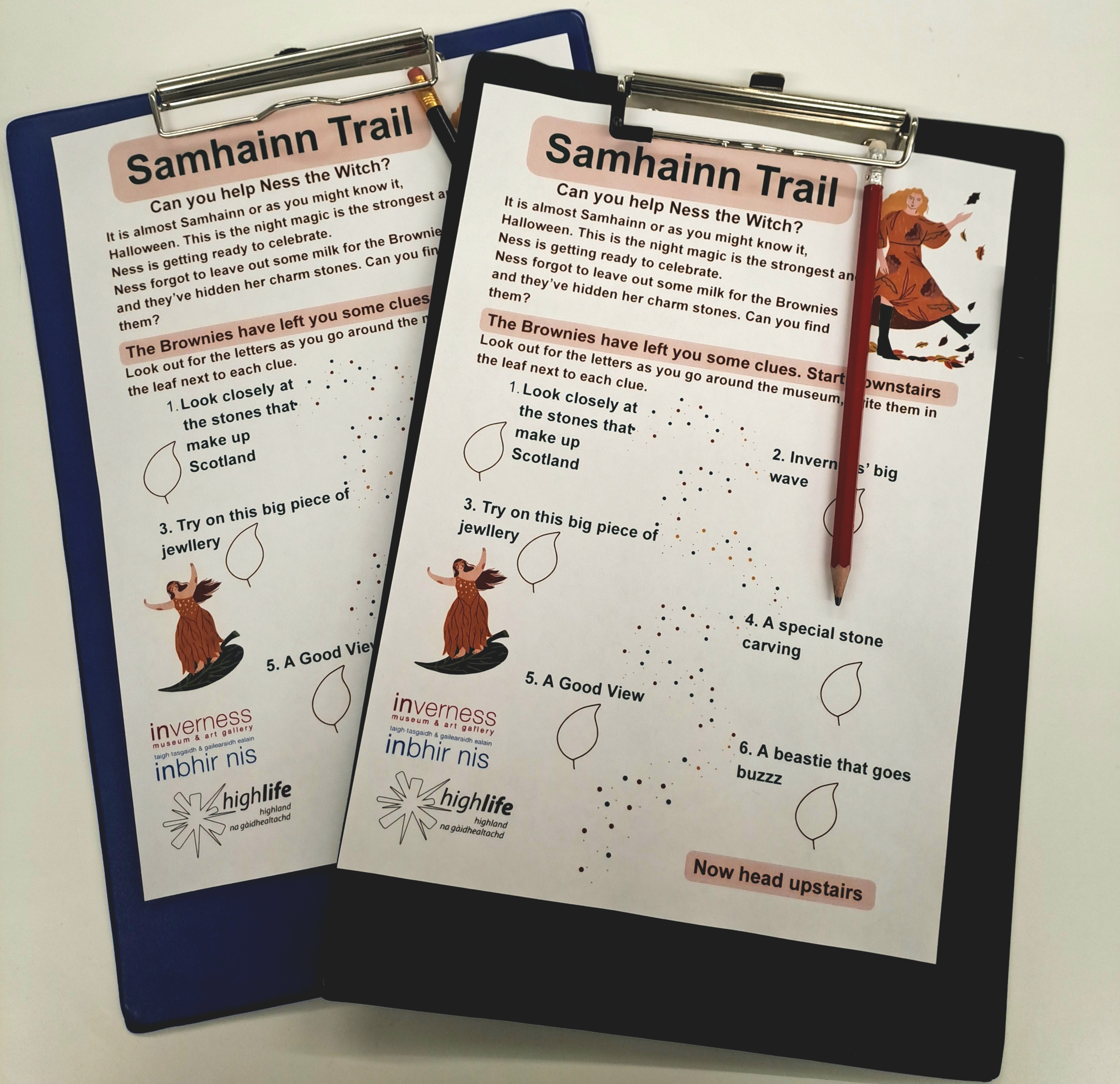 Two clipboards hold “Samhainn Trail” activity sheets featuring a witch illustration, leaf icons, and clues guiding children around a museum. One clipboard is blue and the other black, and a red pencil is clipped to the top sheet.