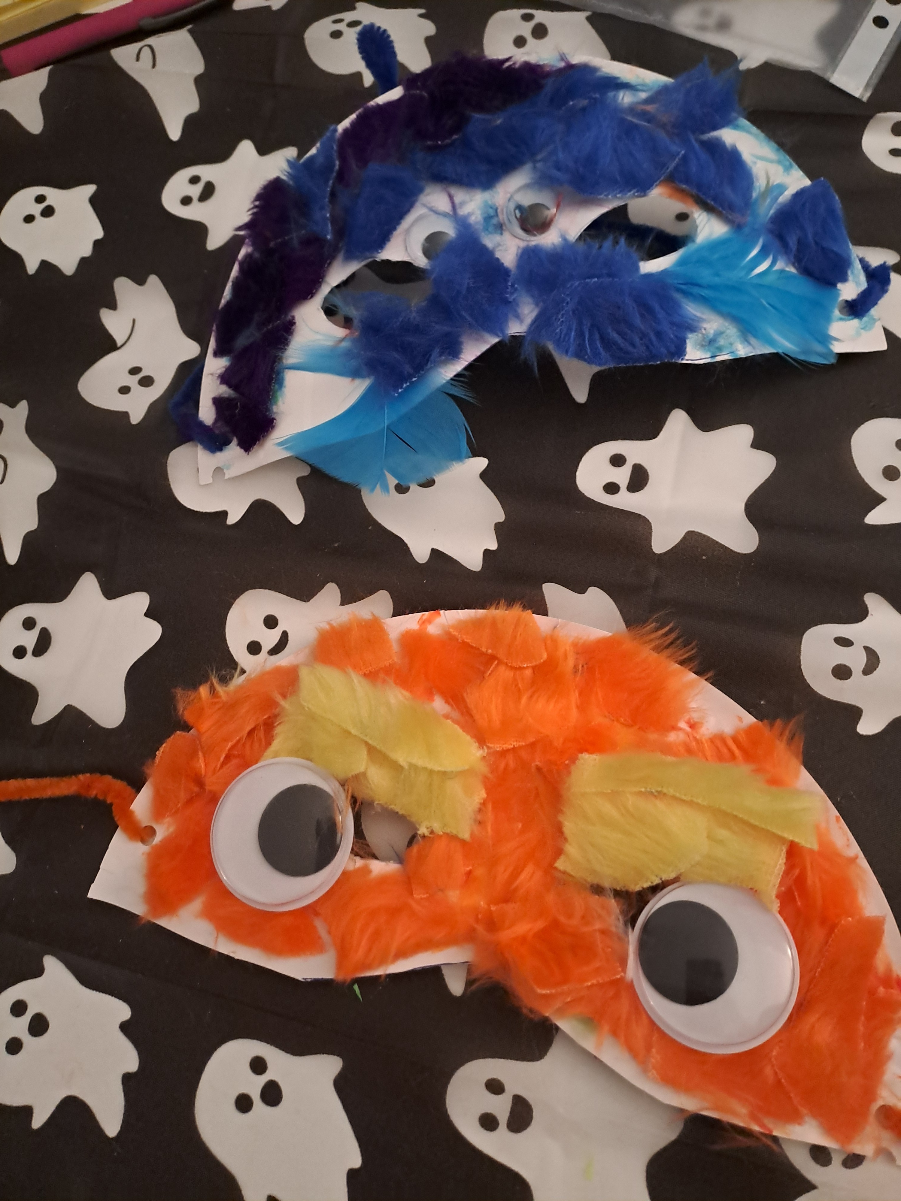 Two colourful handmade masks decorated with feathers and large googly eyes lie on a ghost‑patterned tablecloth—one in orange and yellow, the other in blue and purple.