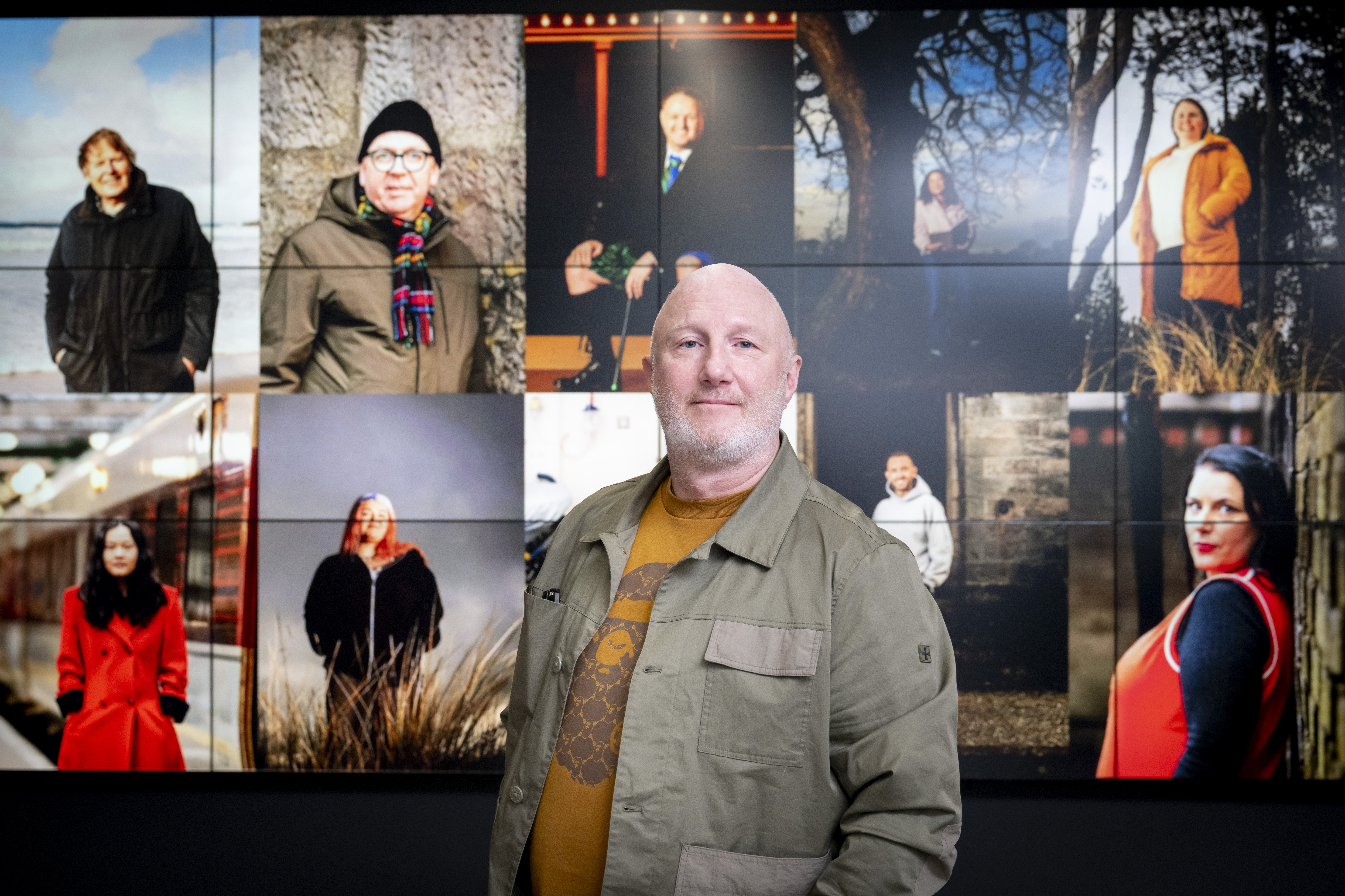 A person standing in front of a large wall display showing a grid of portrait photographs of different people in varied settings.