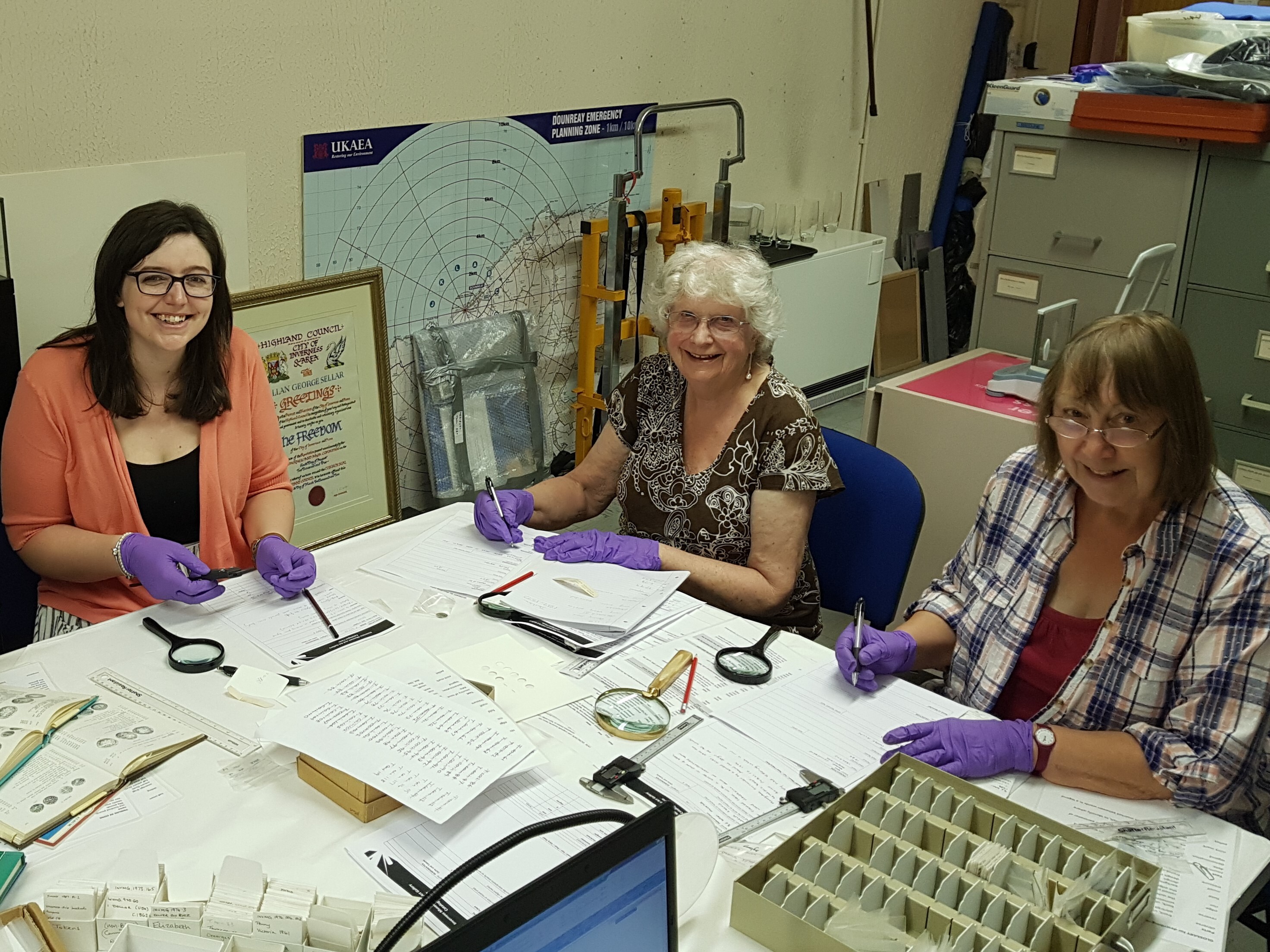 Three people seated at a table sorting and recording coin‑related items using magnifying glasses, tools, and paperwork in a workspace filled with maps, folders, and storage drawers.