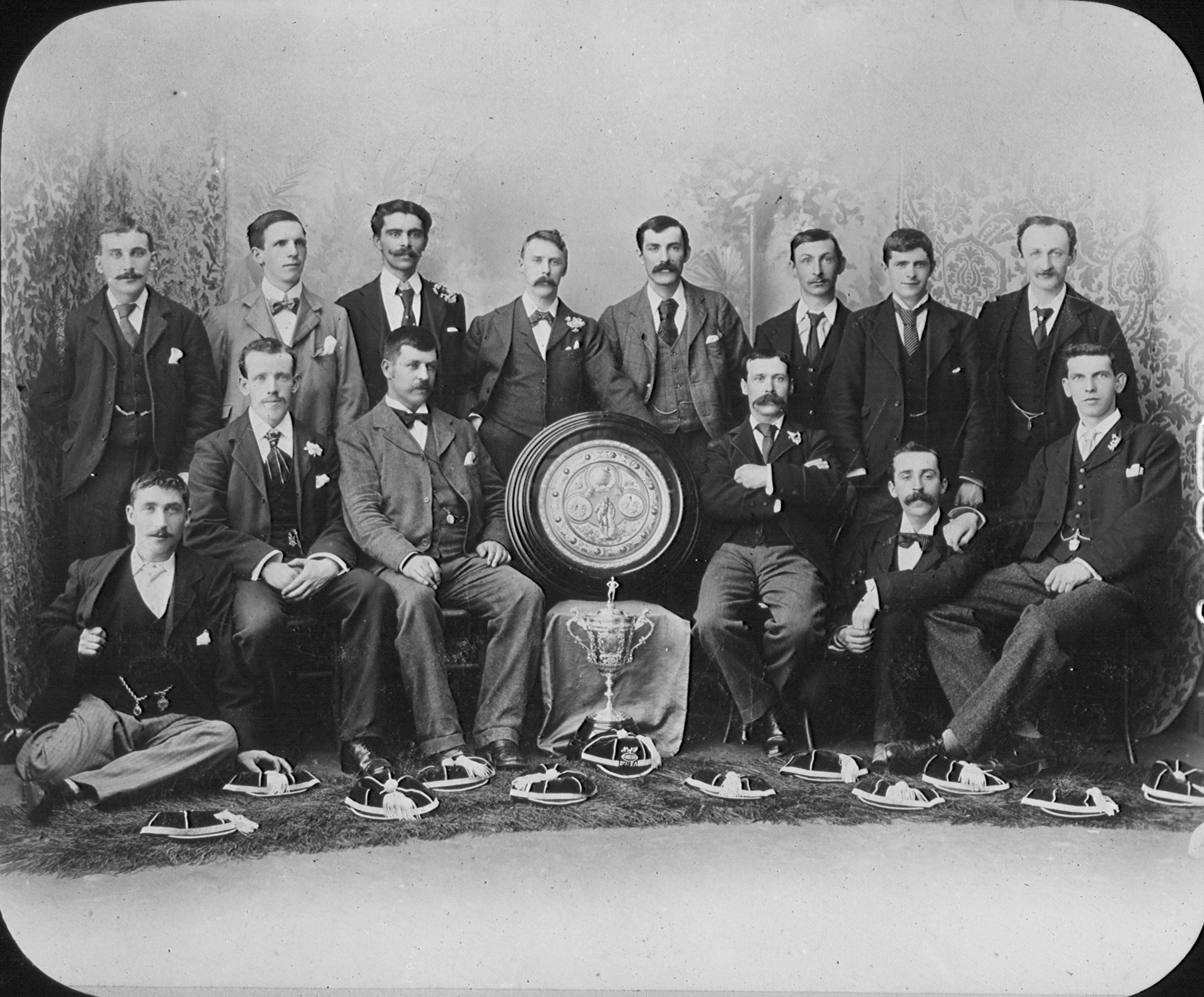 Black-and-white group portrait of a late-19th-century sports team in formal suits, posed with a large shield and trophy, with caps laid out in front.