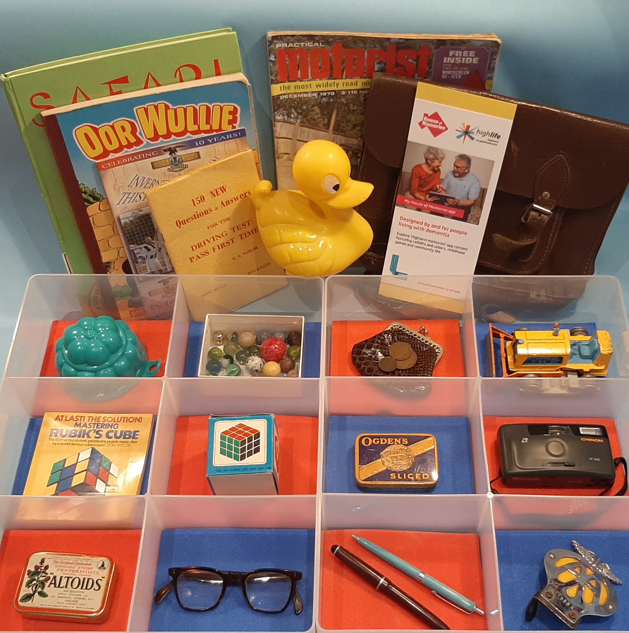 A display of nostalgic items including books, a yellow rubber duck, a leather satchel, marbles, coins, a toy car, a Rubik’s Cube, a camera, eyeglasses, pens, and small tins.
