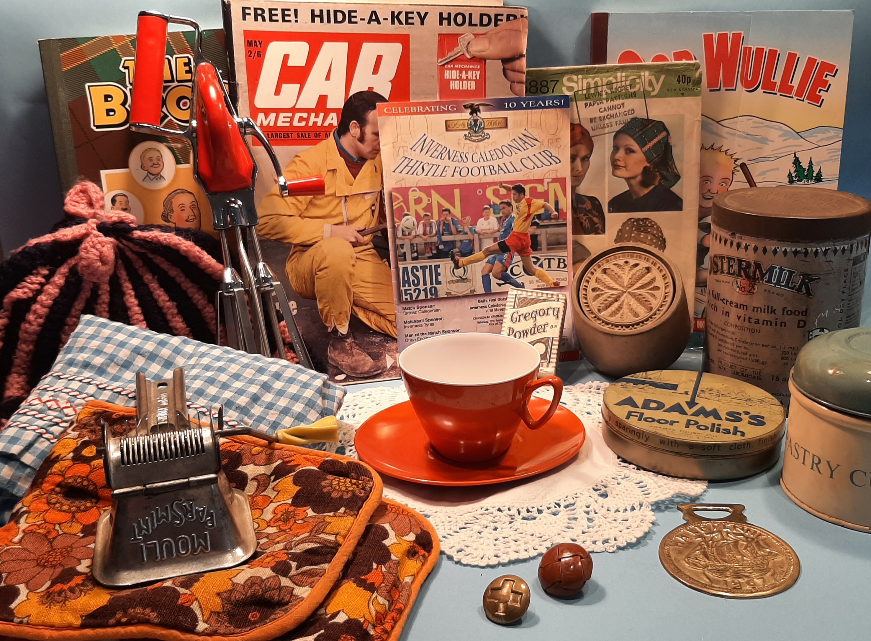 A display of vintage household items and memorabilia, including old magazines, sewing patterns, a red-and-white teacup, kitchen tools, tins, a medal, patterned fabric, and small ornaments arranged on a lace mat.