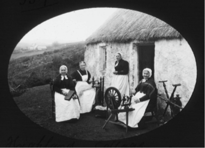 A historical black and white image of four people sitting outside a thatched stone cottage with spinning wheels and textile tools.