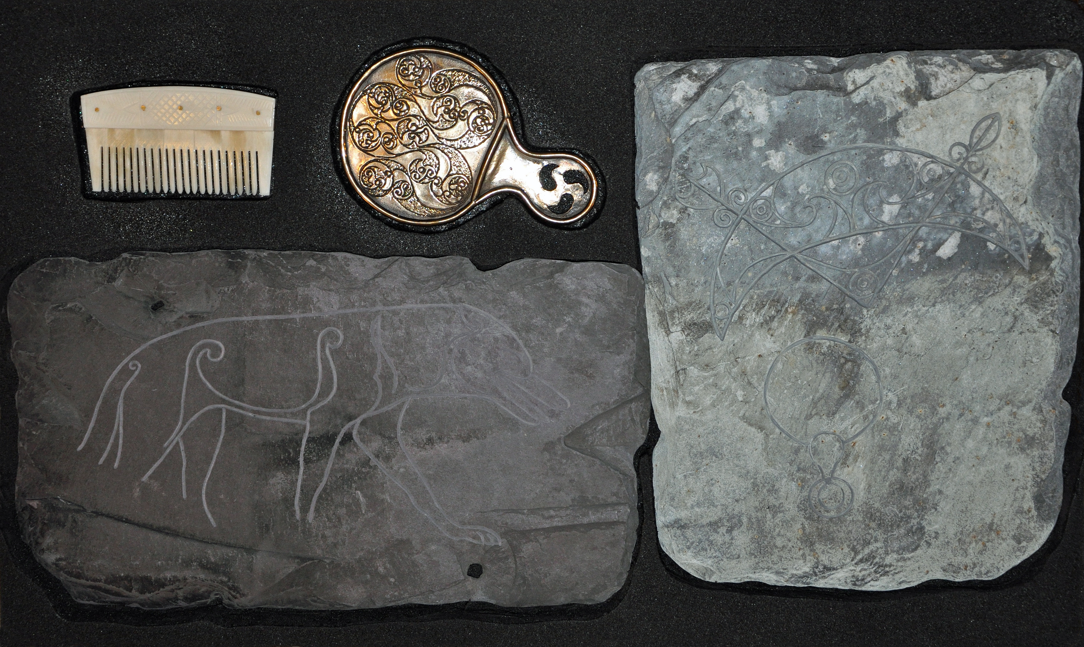 Two stone slabs with carved designs, one white comb and the back of a round metal mirror with intricate inscriptions, all placed on a black background.