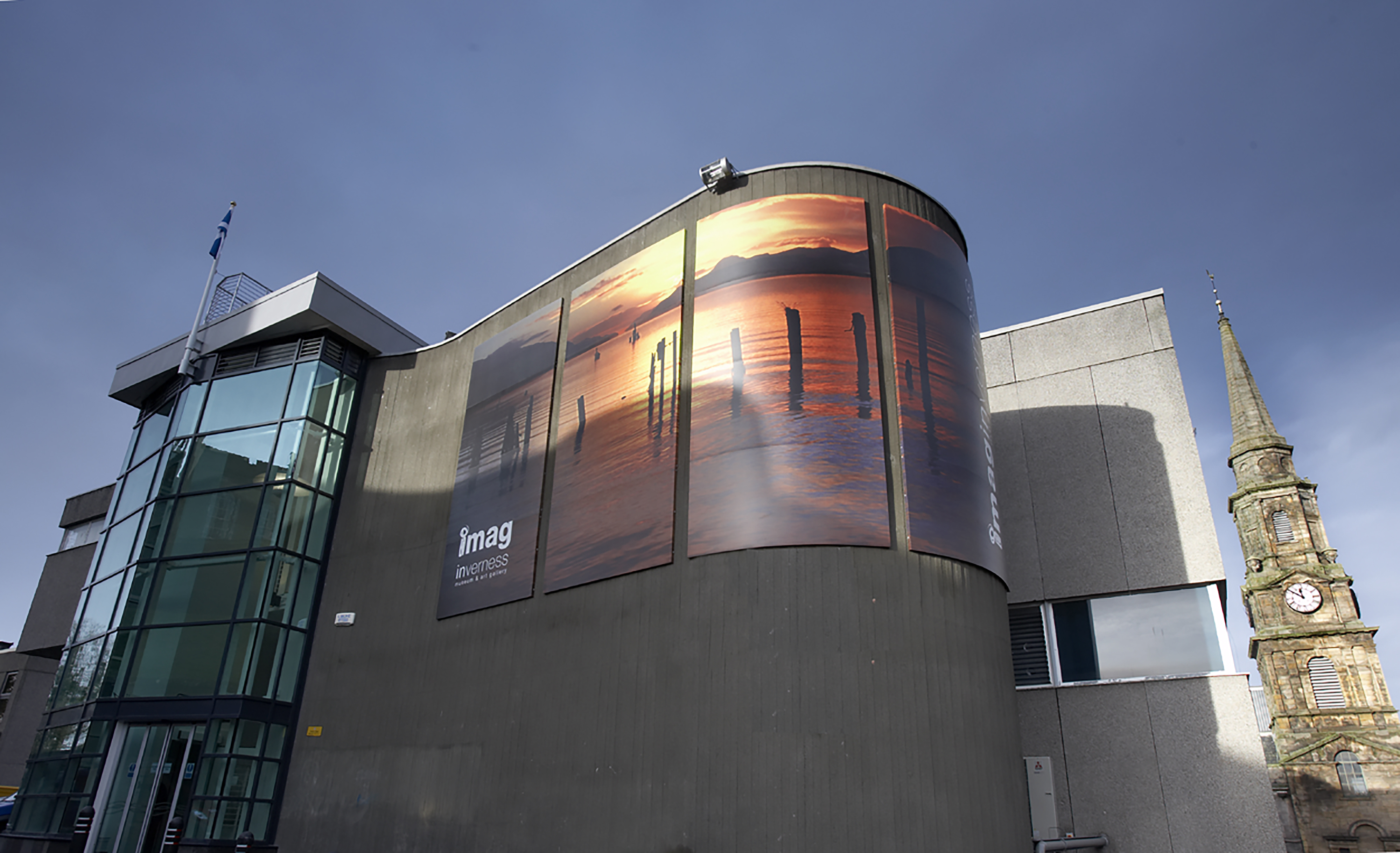 The exterior curve of a concrete building with a glass entrance and older clock tower in the background.