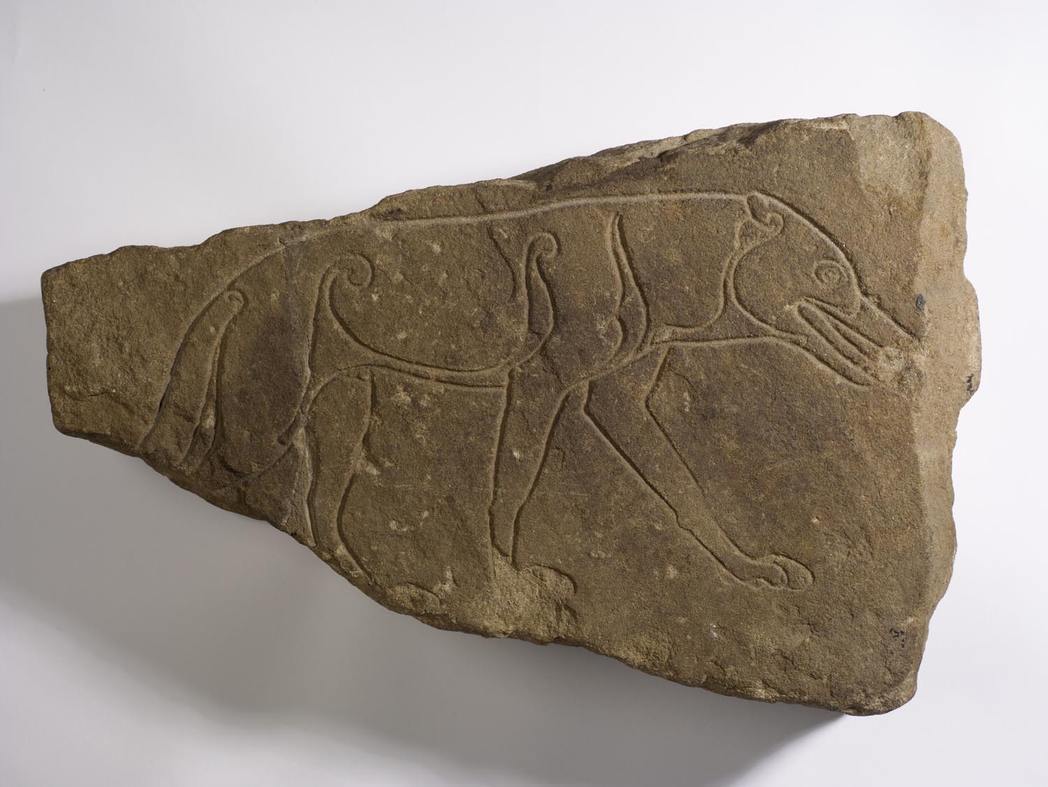 A large, triangular shaped stone with a smooth, brown surface.  A line drawing of a wolf has been carved into the surface.  One of the back legs and the end of the tail are missing.