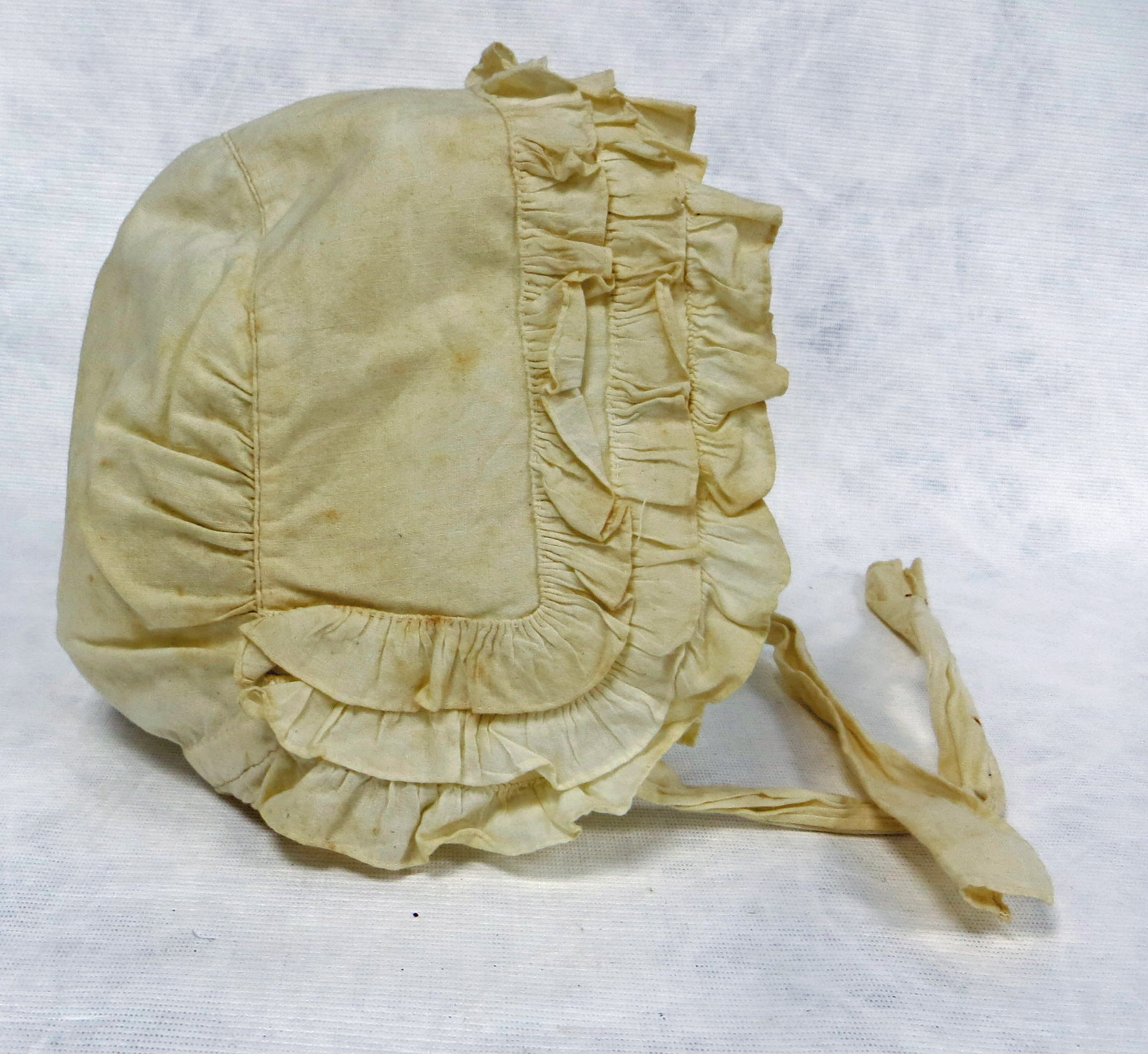 A cotton mutch, an old-fashioned type of bonnet with a frilled edge, worn by ladies. The bonnet should be white but is stained yellow.  Shown before conservation work.