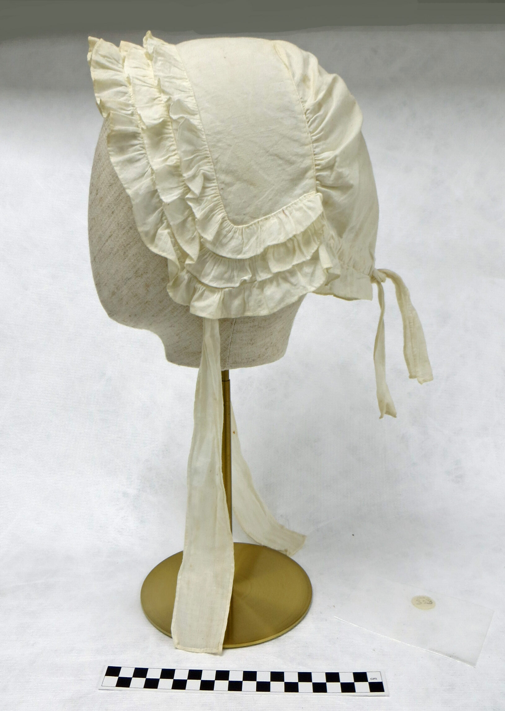 A cotton mutch, an old-fashioned type of bonnet with frilled edge, worn by ladies.  Mounted on a display head stand. Shown after conservation work, the bonnet is white, clean and ironed.