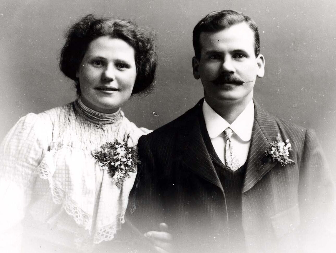 A black and white photograph of an Edwardian couple against a plain backdrop, shown from the waist up.  The lady, on the left, wears a white blouse with a frilled collar and her hair is pinned up.  The gentleman, on the right wears a suit, shirt and tie, and has a waxed moustache.  Both are wearing flower corsages on their clothes.  The photo is faded towards the bottom.
