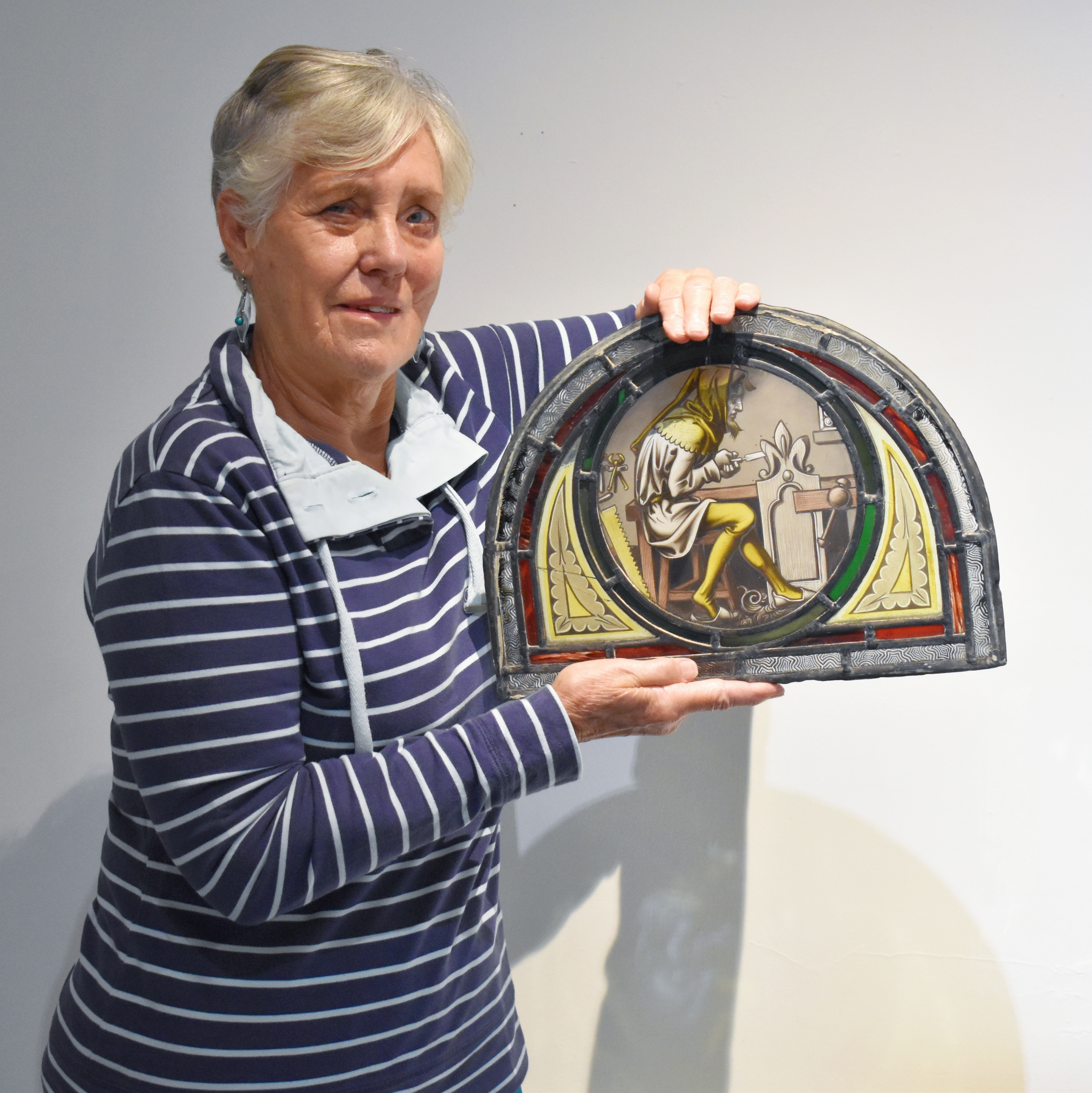 A lady with short hair and a stripey navy top is holding a semi-circular stained glass panel.  The panel depicts a woodcarver in medieval dress, in yellow tones with decorative borders in green, yellow and red glass.
