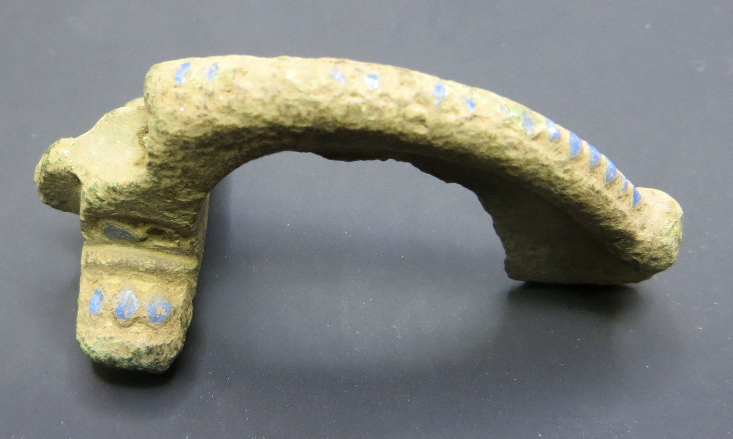 A Roman fibula brooch which is a long, arched type of metal brooch. It is missing its pin. Shown before conservation when it is caked in pale earth.