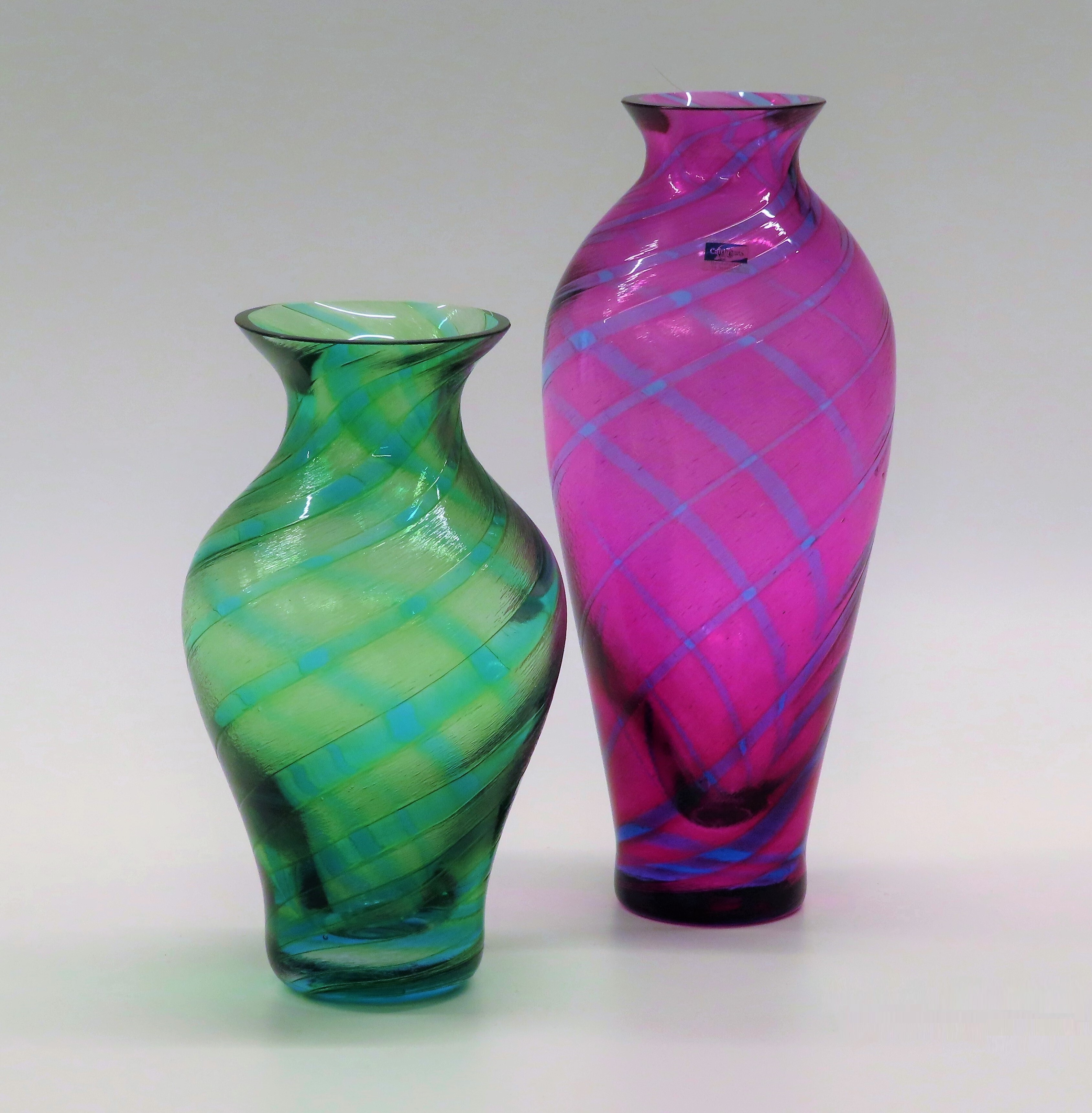 Two vases on a white background.  On the left, a shorter green vase with blue stripes, and the right a tall deep pink vase with blue stripes.  The stripes run diagonally in both directions, giving the appearance of tartan.
