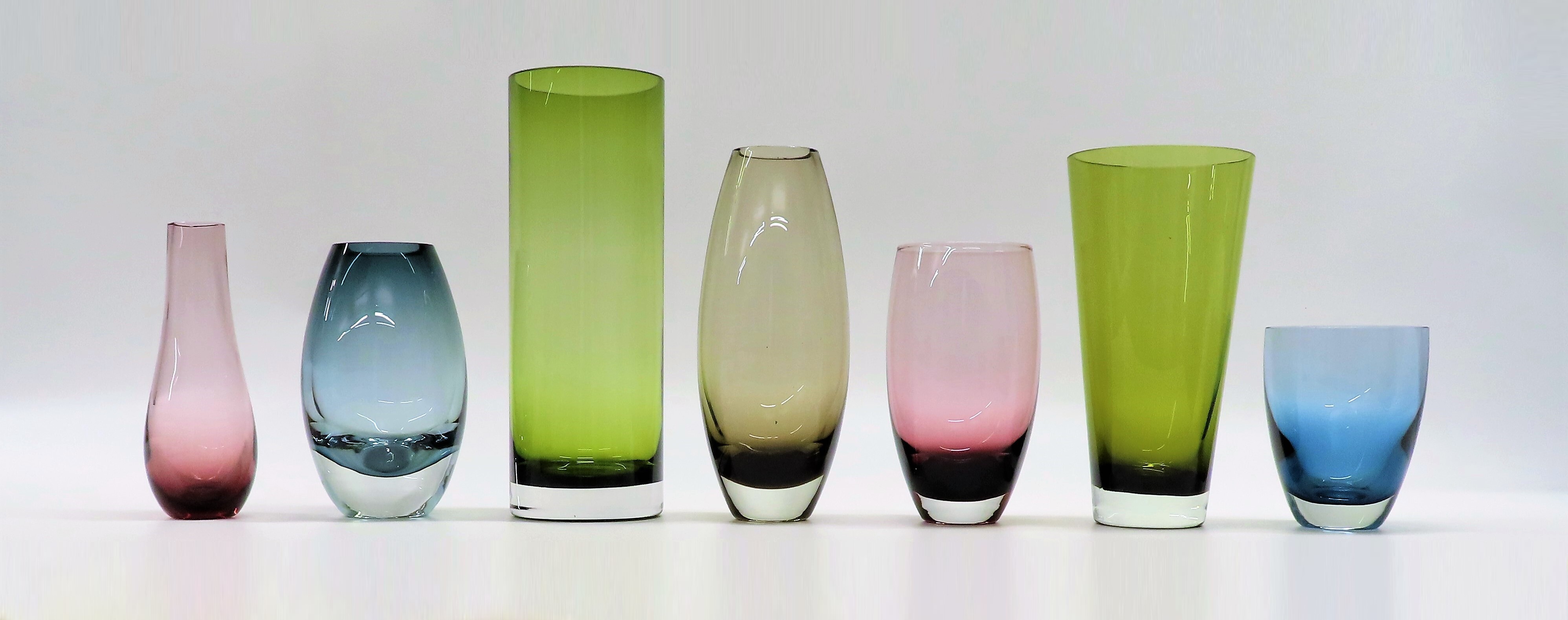 A row of coloured glass vases of different shapes, against a plain white background.  The row replicates the row of vases seen in a vintage Caithness Glass postcard.
