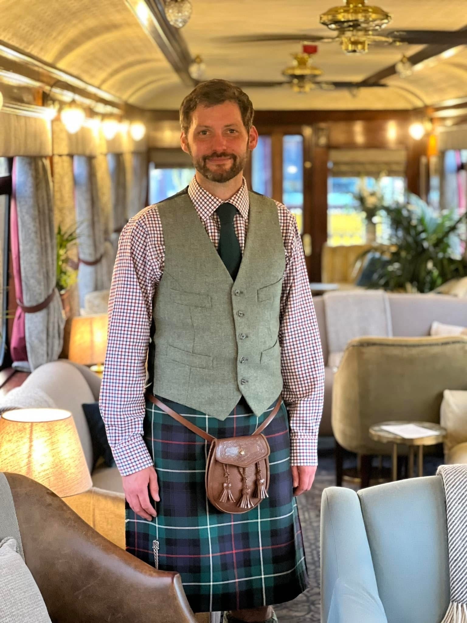 Portrait photograph of historian and tour guide, Alexander Grant McKenzie, wearing Highland dress and standing aboard a luxury train carriage.  Image supplied by Alexander Grant McKenzie.