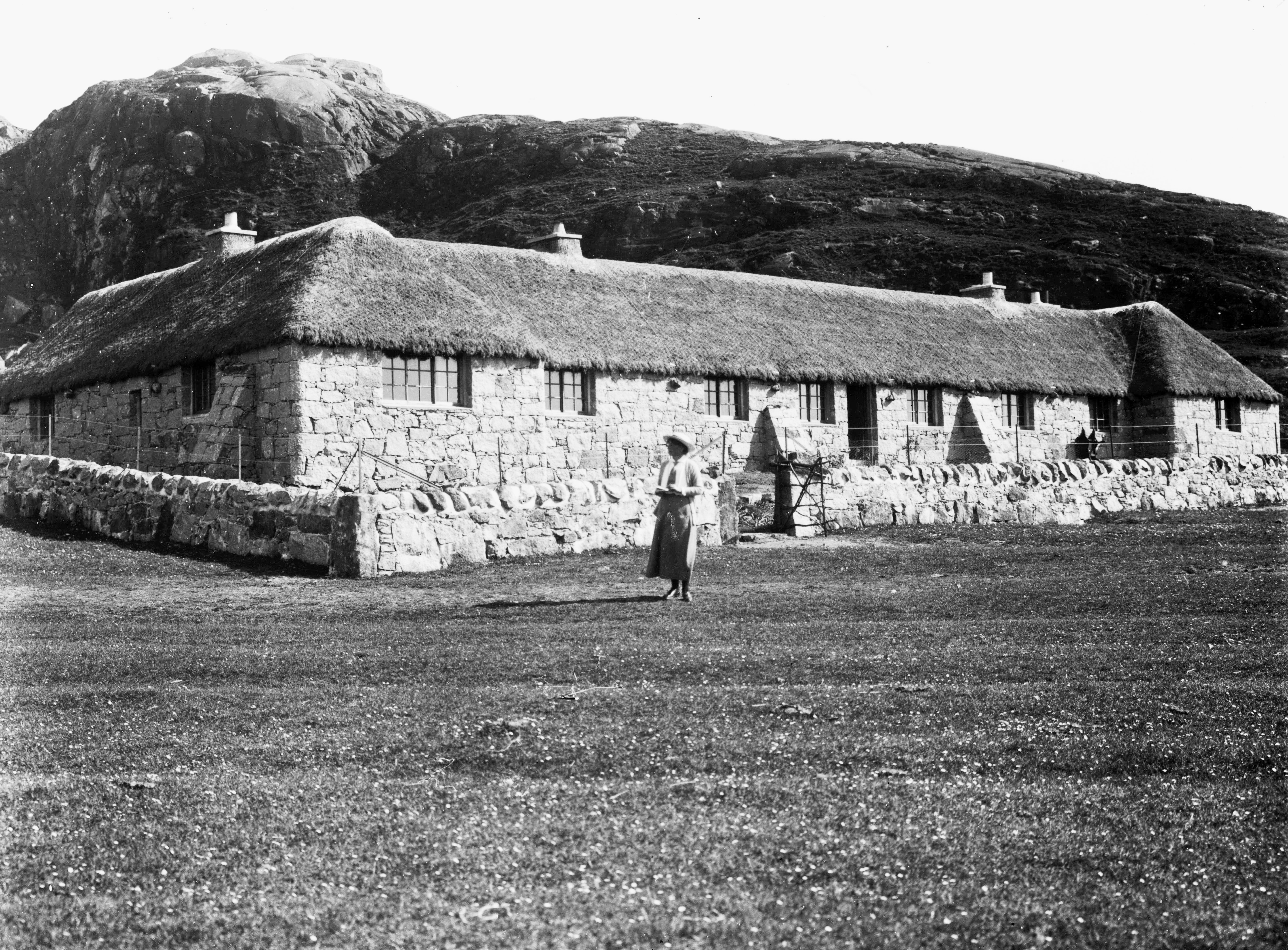 A black and white photo of a long, low building with stone walls, small windows and thatched roof. A rugged hill rises from behind the building. A lone female figure stands in front of the building, looking away to the left.