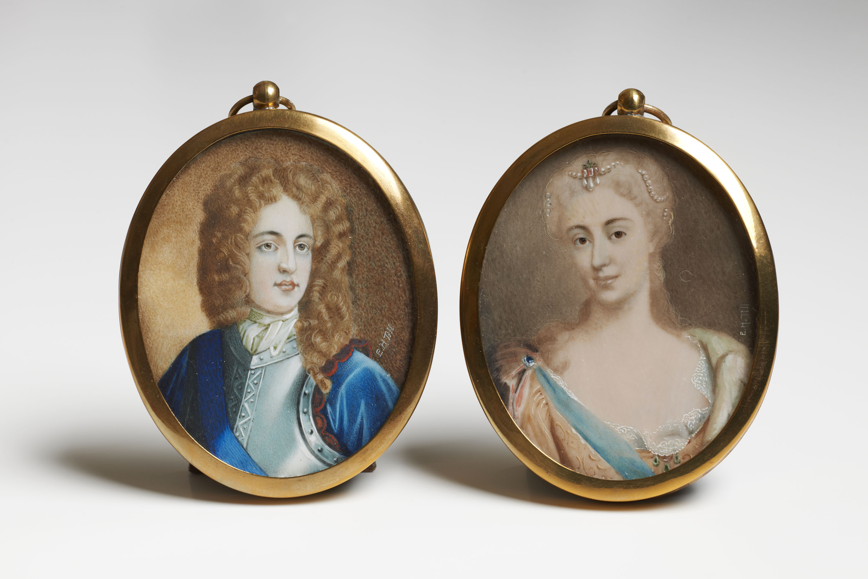 Two oval miniature portraits in matching, plain gold frames. On the left is a man wearing a blue velvet jacket and a long wig of brown curls.  On the right is a lady wearing pearls in her hair and a lacey dress with a low scoop neckline.  These are the parents of Charles Edward Stuart (Bonnie Prince Charlie).