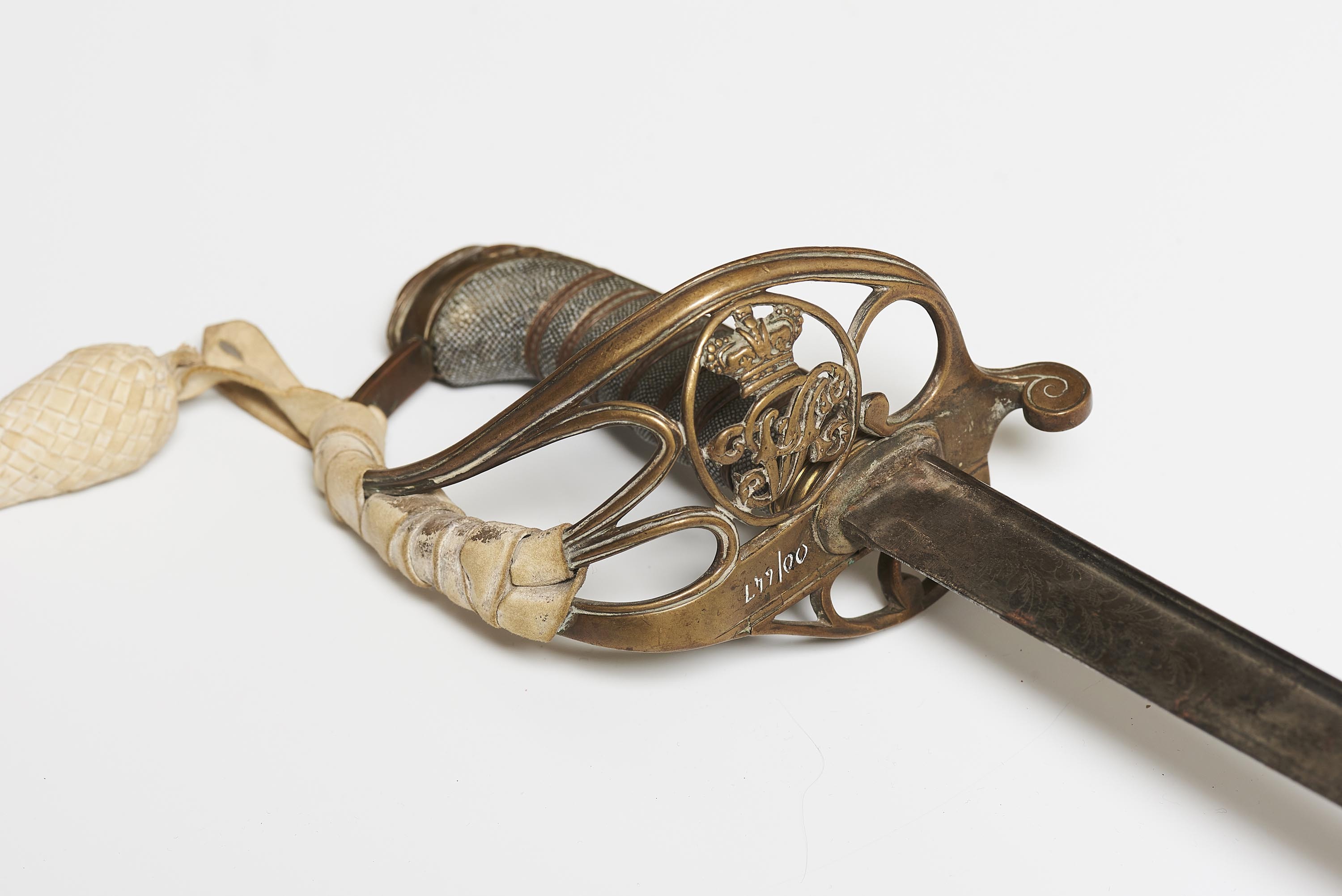 Close-up view of a sword hilt, brass metal in a curved design featuring initials VR (for Queen Victoria) and a cream tassle braided onto the handle.