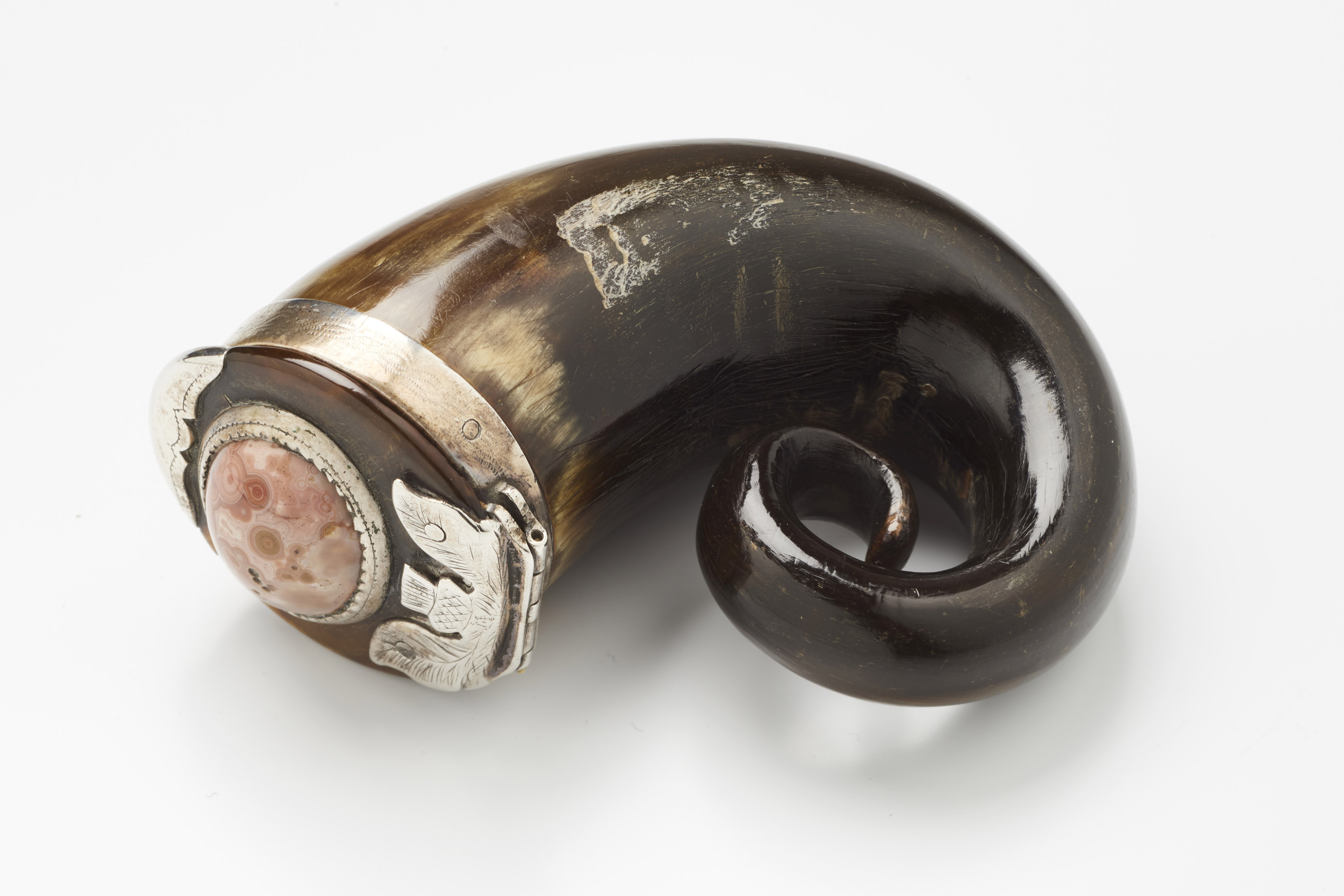 A snuff mull, which is a vessel made from the tip of a cow horn curled up in a spiral, with a lid over the end.  The lid has a silver hinge shaped like a thistle and there is a round jewel of polished pink agate in the centre.