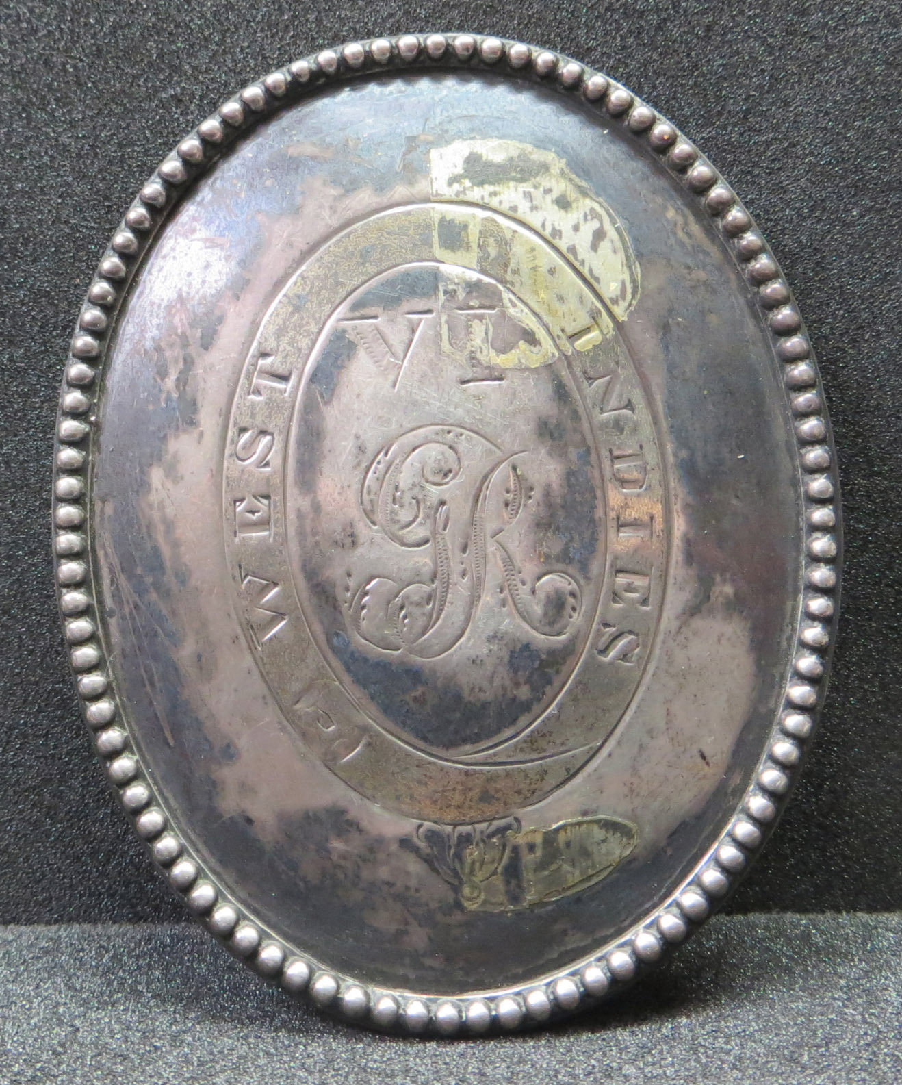 An oval metal badge, worn by a soldier, with a decorated rim and engraving in the middle.  The metal is very tarnished.  Shown before conservation.