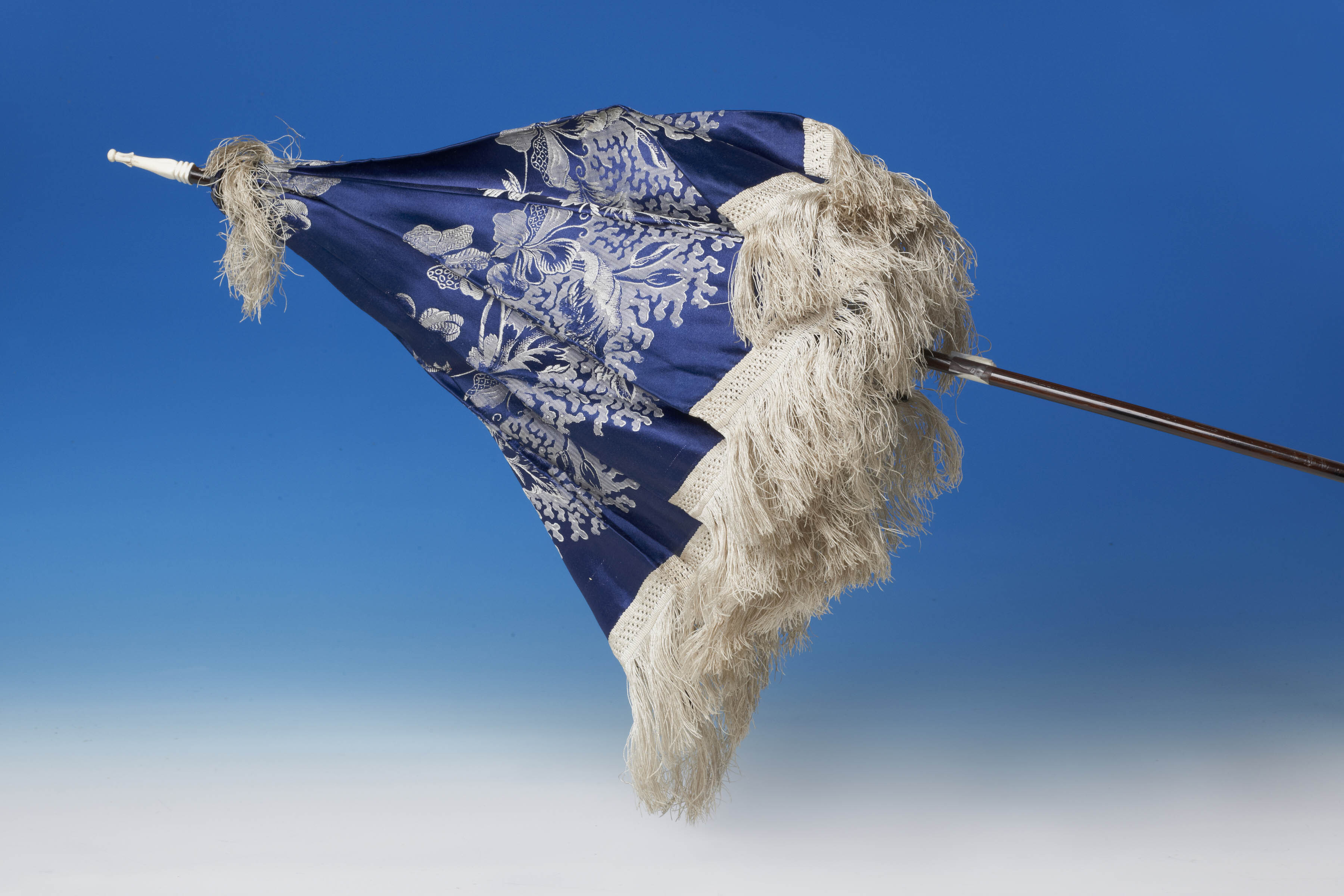 Blue silk parasol with white embroidered floral decoration and white tip.  Shown closed, with tassel trim hanging down around the edge. Set against a blue background.