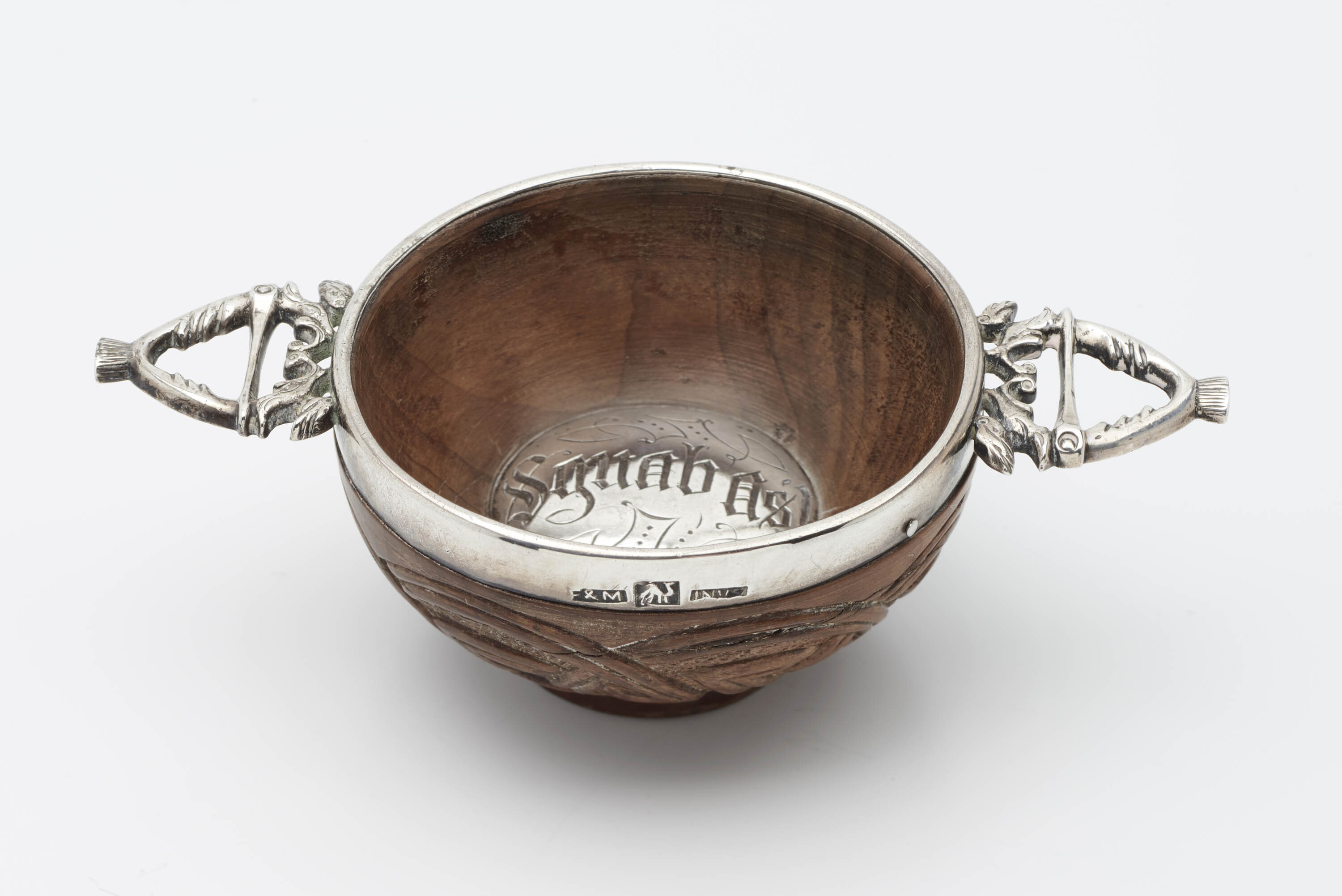 A quaich, consisting of a small carved wooden bowl with a silver rim, two silver heart-shaped handles protruding from the rim, and a silver button in the base engraved with Gaelic text.  A quaich is a traditional two-handled drinking cup from Scotland.