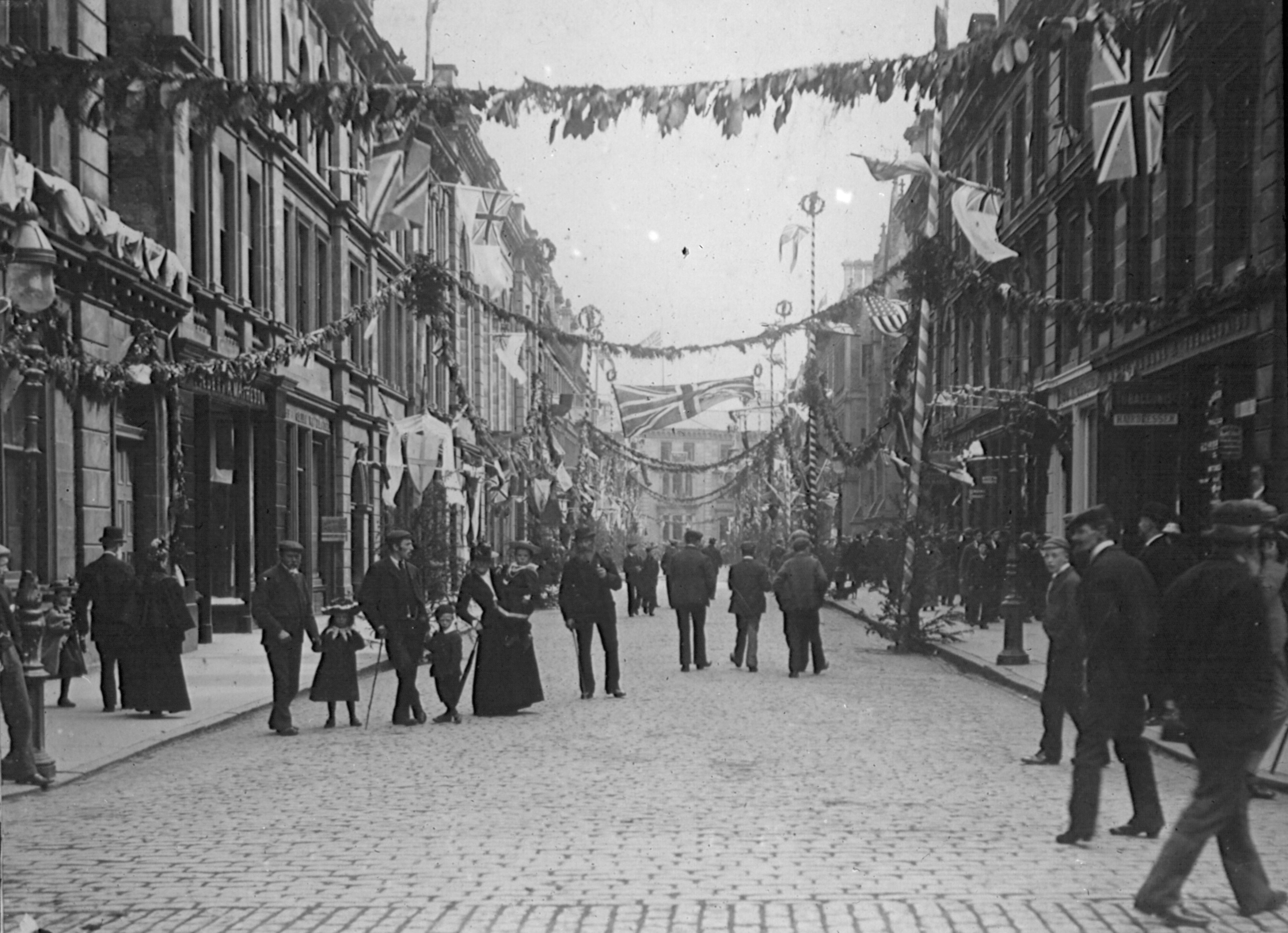 A black and white photograph showing a view down Union Street in Inverness in 1897.  The street is busy with people including families. Above, stings of bunting and flags are hung across the street to celebrate Queen Victoria's Diamond Jubilee.