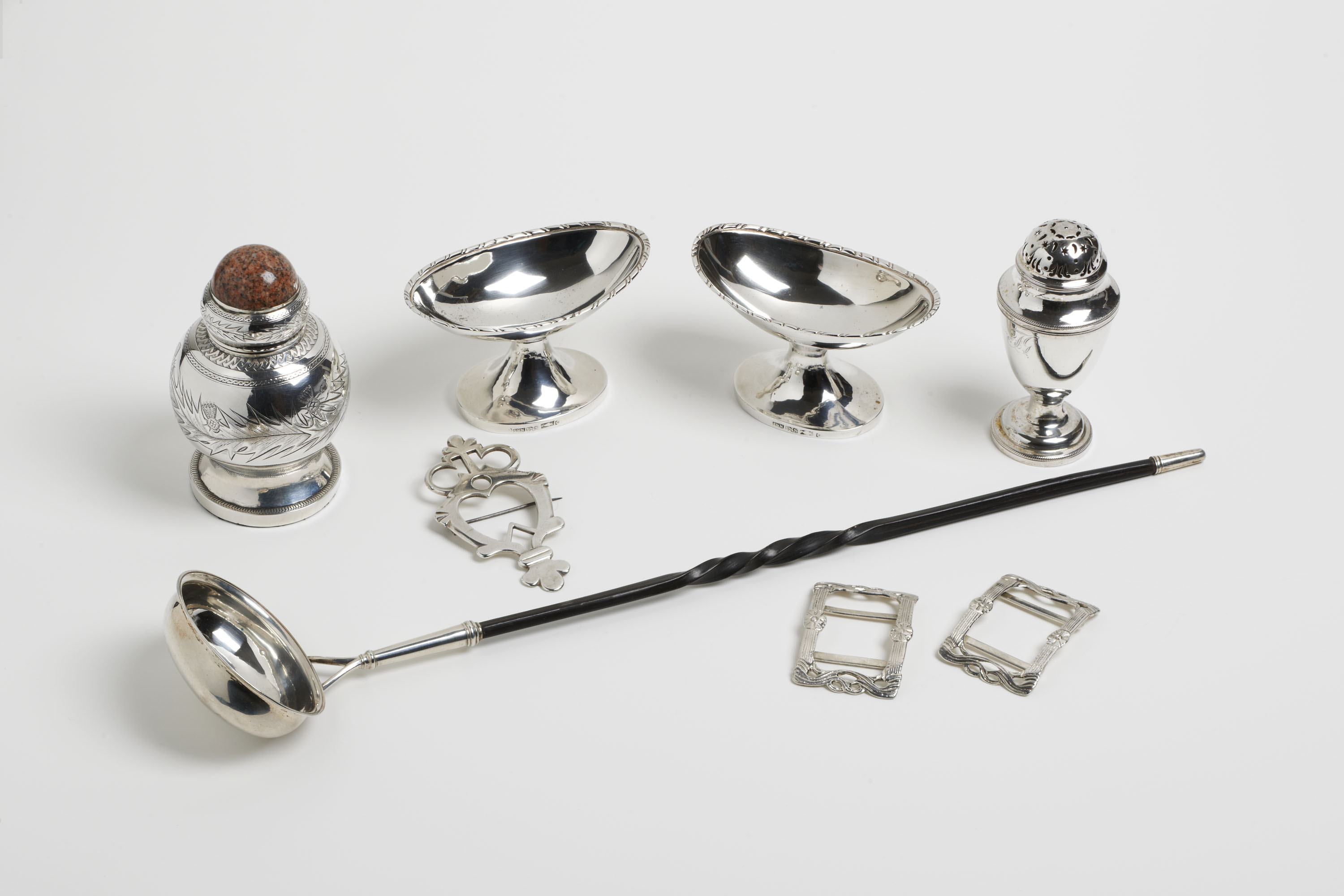 A group of antique silver items, including an inkwell with polished stone lid, a heart brooch, a pair of oval salt dishes, a sugar sifter, a punch ladle with a very long handle and a pair of rectangular shoe buckles. Set against a plain white background.