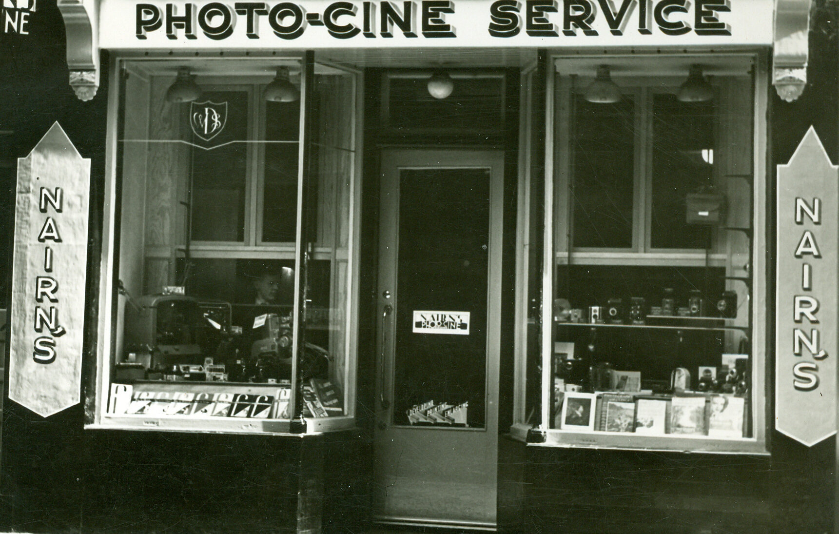 A black and white photograph of a shop front.  There is a central glass-panel door and two large windows either side with displays of cameras and photographs.  The shop sign above says Photo-Cine Service and the name Nairn's appears on the door and on the walls either side of the windows.