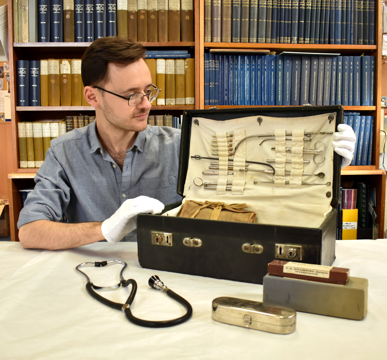A young man wearing a blue shirt and white cotton gloves is sitting at a desk.  He is looking at a doctor's case and holding the lid open to reveal medical equipment inside.  A stethescope and some medical tins sit on the desk.  Behind him is a bookcase full of old books.