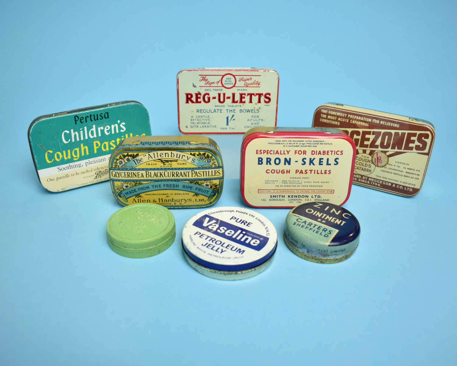 8 vintage tins of medicine against a blue background.  The tins are brightly coloured and include vaseline, children's cough pastilles, zinc ointment and reg-u-letts.