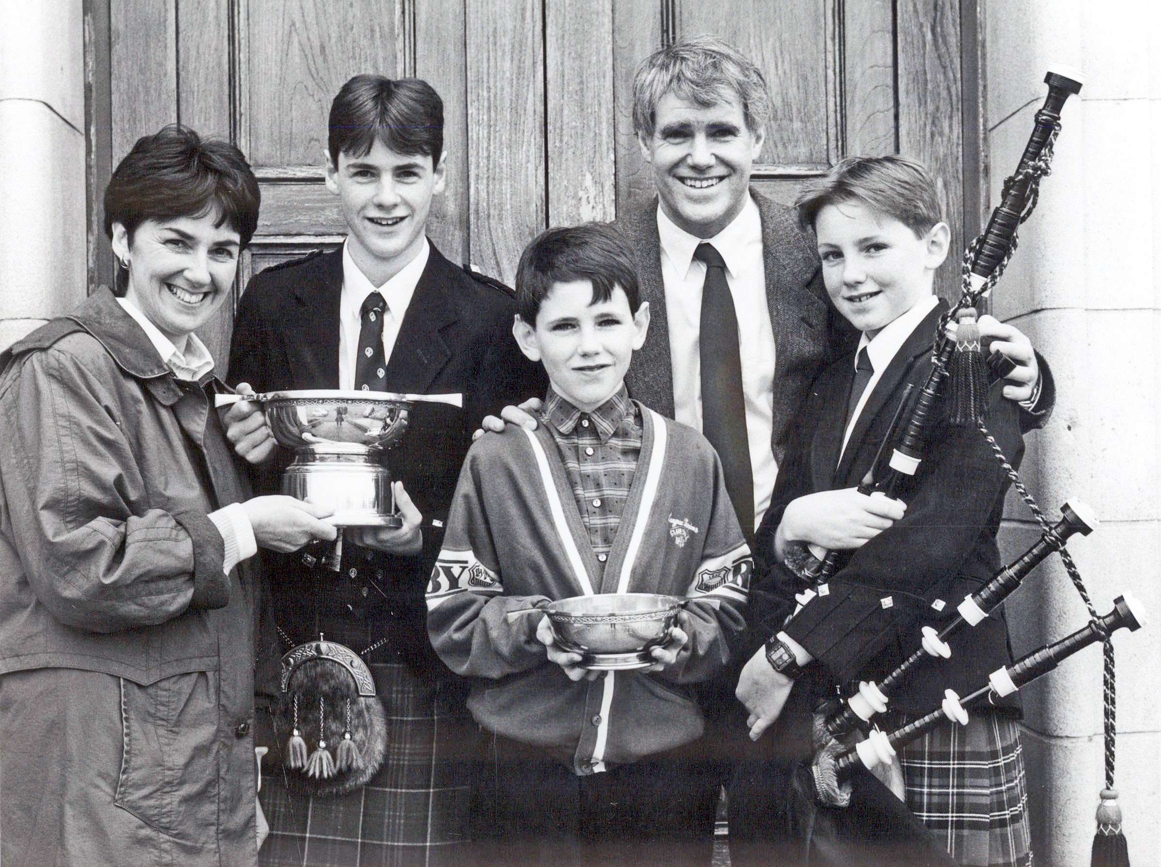 A black and white photograph of a family of five people (mother, father and three sons) standing in front of a pair of wooden doors.  Two of the boys are holding quaich trophies and one boy is holding a set of bagpipes. All of them are smiling at the photographer.