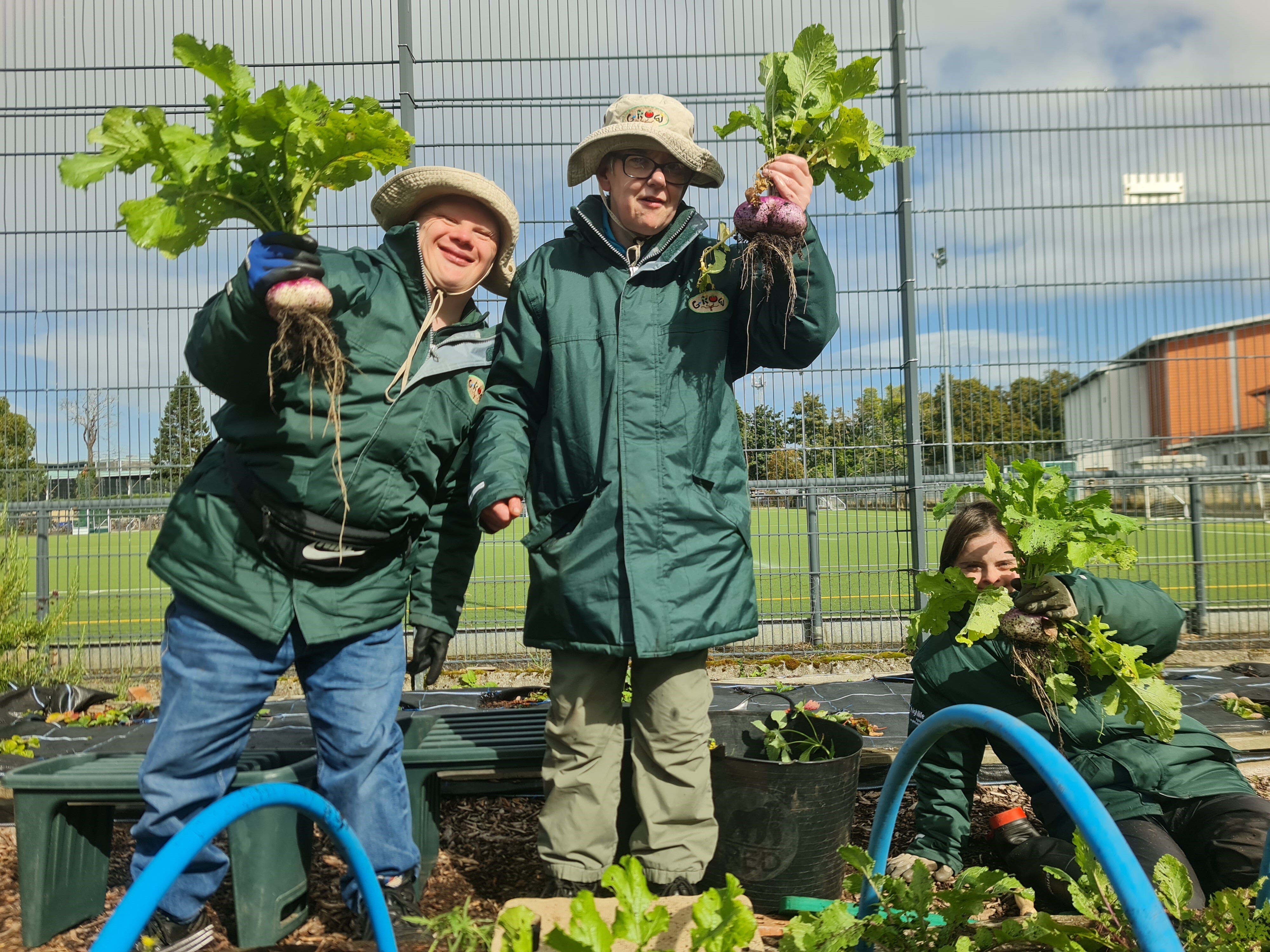 Three people wearing green gardening jackets and sun hats proudly hold up freshly harvested root vegetables in an outdoor garden area. They stand and kneel among raised beds, with green hoops, mulch, and plants visible around them. A sports field and tall metal fencing are in the background under a partly cloudy sky.