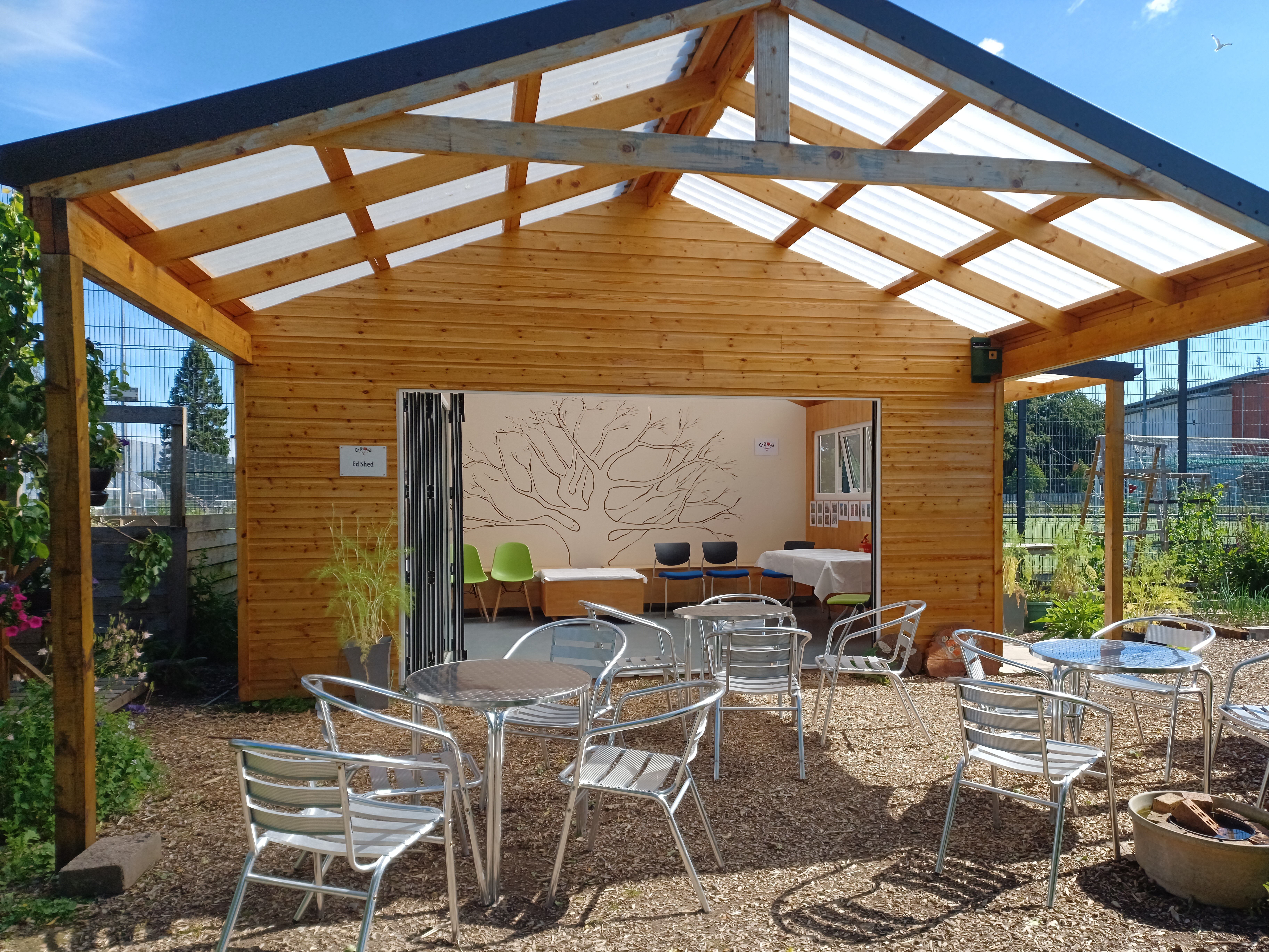 Patio doors wide open from an education shed with tables and chairs set up under the porch on a beautifully sunny day.