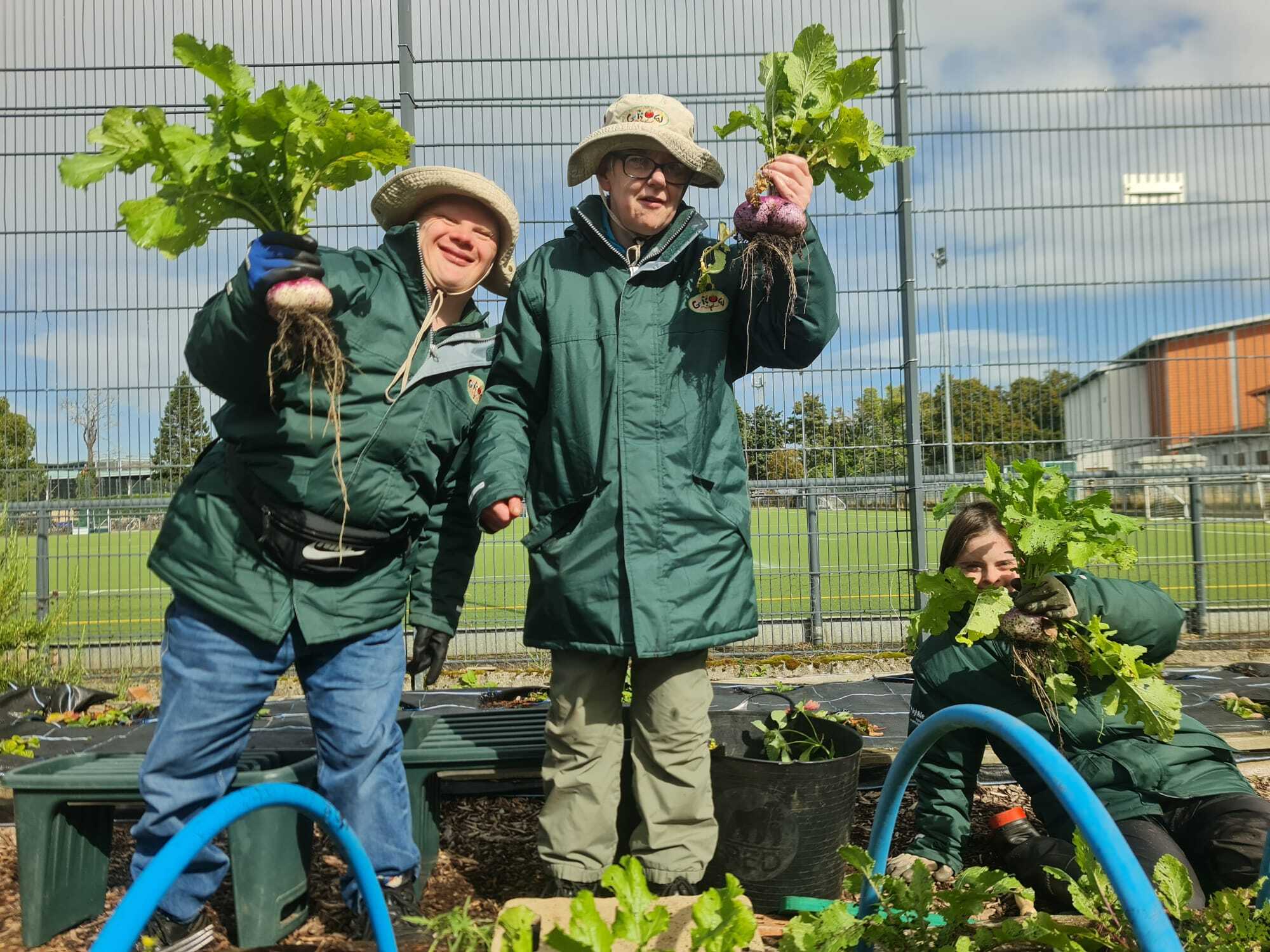 Three people wearing green gardening jackets and sun hats proudly hold up freshly harvested root vegetables in an outdoor garden area. They stand and kneel among raised beds, with green hoops, mulch, and plants visible around them. A sports field and tall metal fencing are in the background under a partly cloudy sky.
