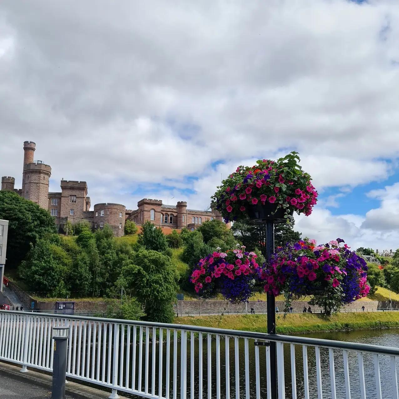 Colourful hanging baskets along a bridge with the view of Inverness Castle in the background.