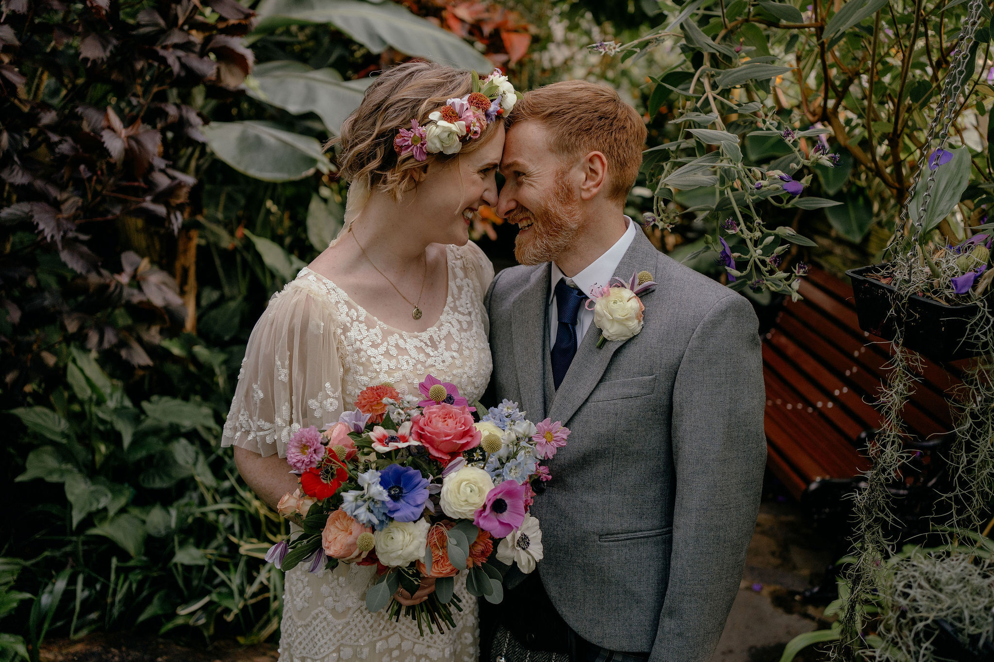 A wedding couple stands close together in a lush indoor garden setting. The person on the left wears an ivory dress with intricate lace detailing and sheer sleeves, accessorized with a floral crown and a pendant necklace. They hold a vibrant bouquet featuring roses, ranunculus, and other flowers in shades of red, pink, purple, blue, and cream, accented with greenery. The person on the right wears a grey suit jacket with a white shirt, navy tie, and a boutonnière of a white rose with purple accents. Behind them is a wooden bench surrounded by dense green foliage and hanging plants with small purple flowers.