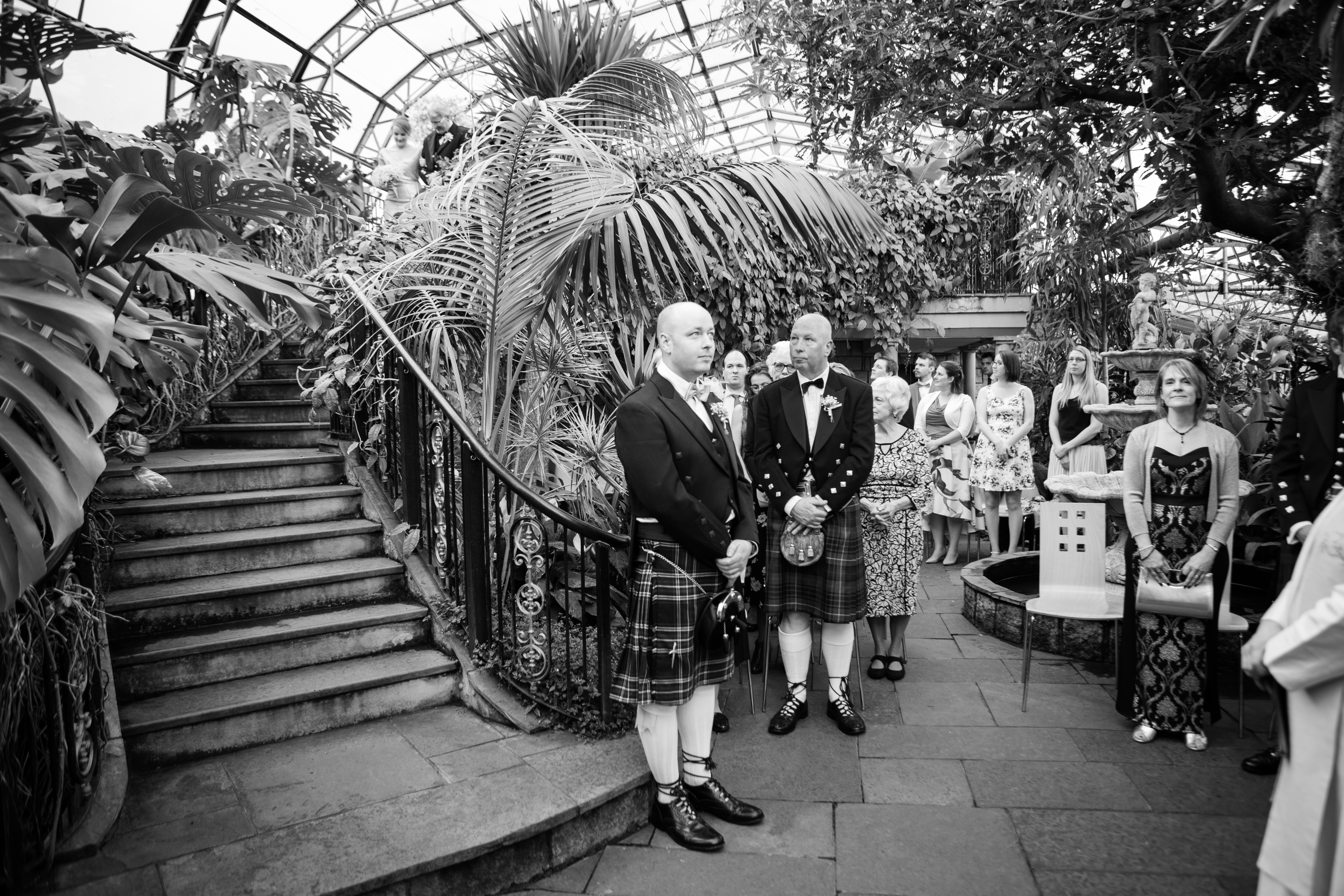 A black-and-white photo of a wedding ceremony inside a glasshouse filled with lush tropical plants.