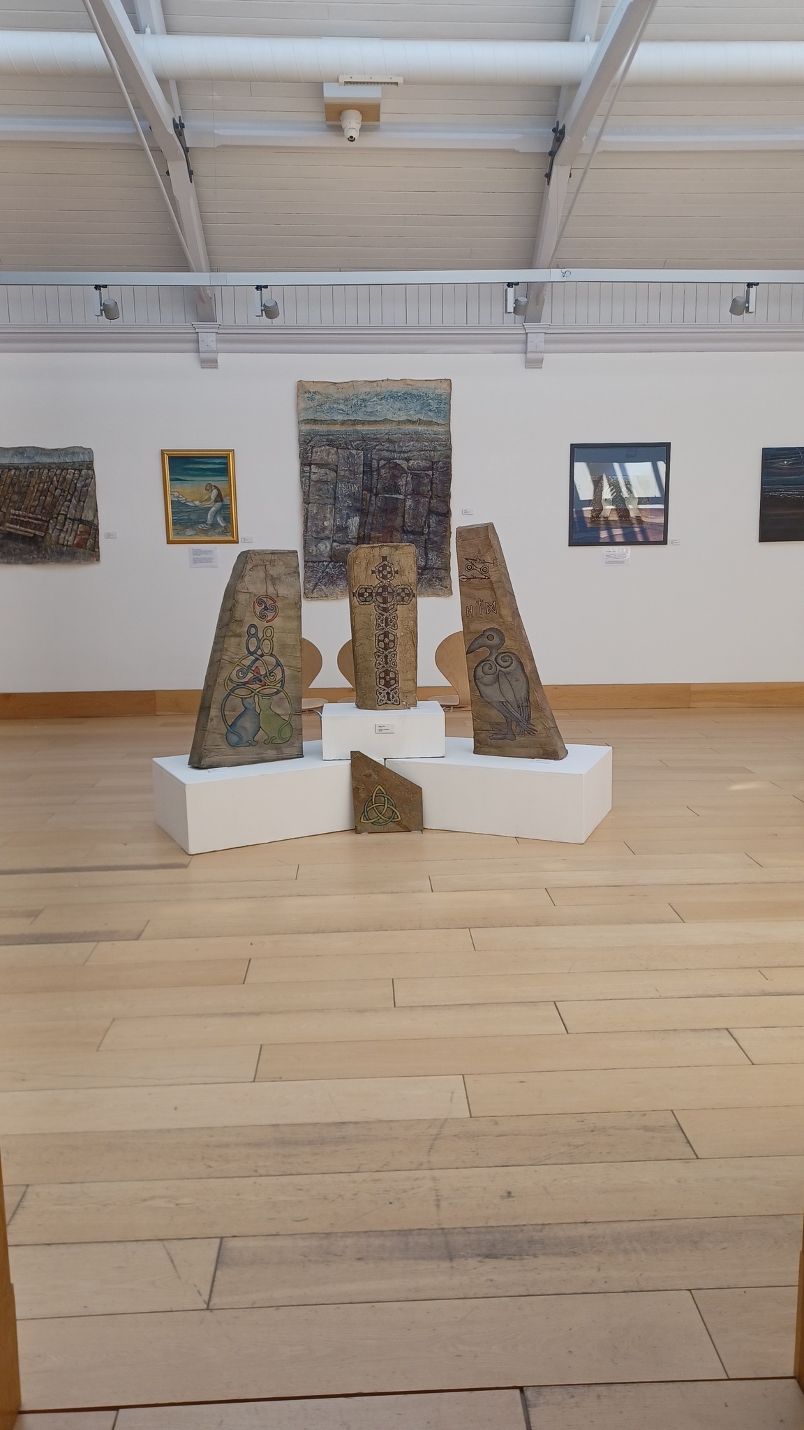 Gallery display featuring four carved stone pieces with Pictish-style designs arranged on white stands, surrounded by framed artworks on a light-colored wooden floor.